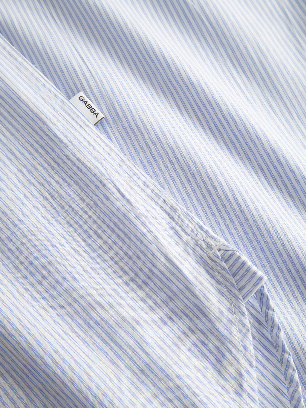 A close-up of the white and light blue pinstripe fabric, labeled with Gabbas Sven Reg. Form LS - White Stripe, is crafted from 100% cotton. The shirt features neatly stitched seams, embodying the quality expected from a premium brand.