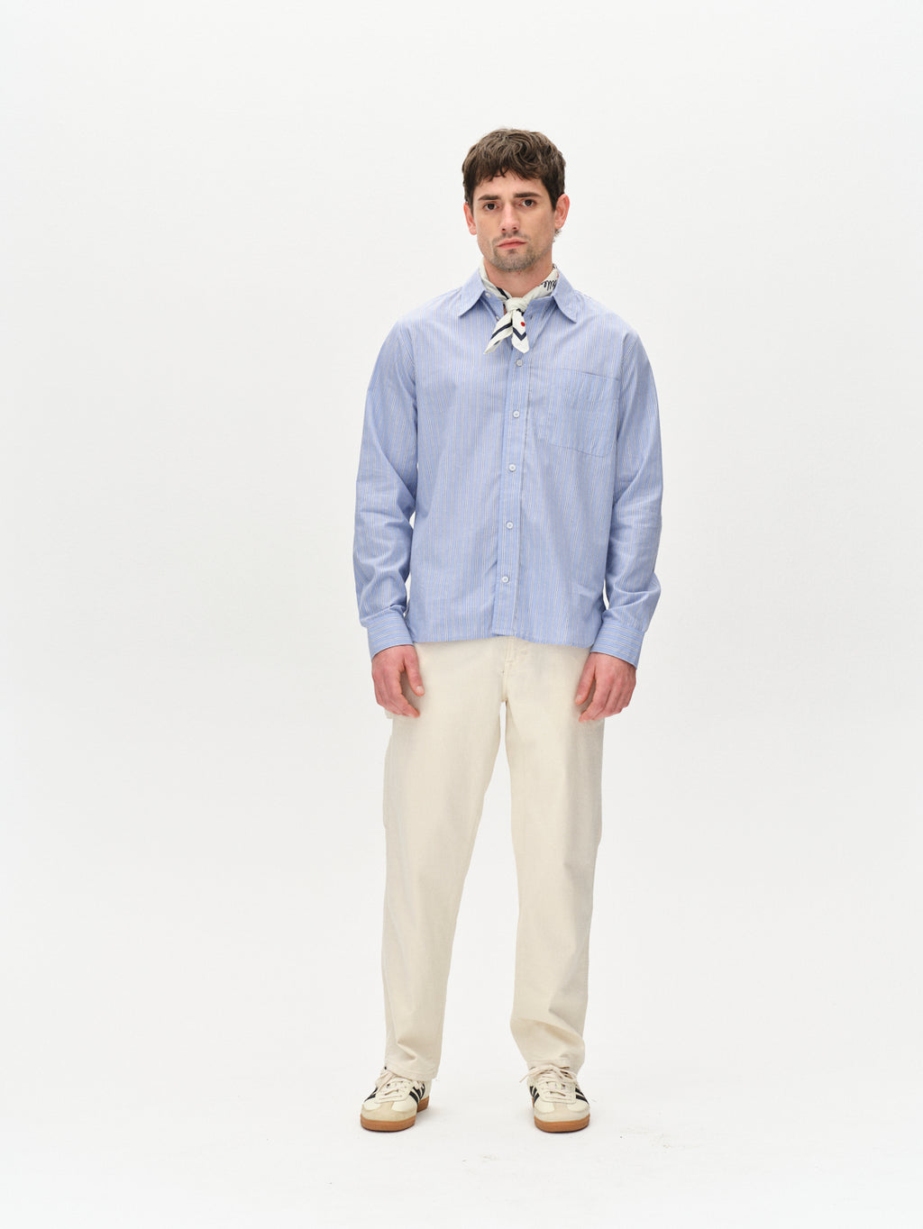 A man with short brown hair stands against a plain white background, wearing the Gabba Sven Reg. Form LS - Blue Stripe shirt, beige pants, and white sneakers while looking directly at the camera with his hands by his sides.
