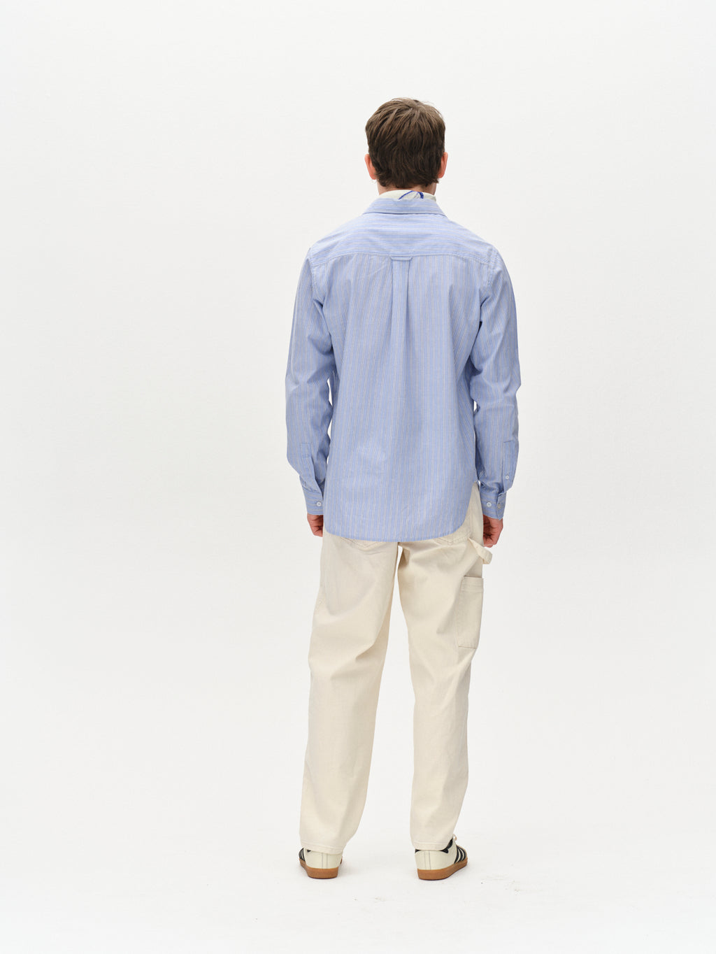 Facing away, the person wears Gabbas Sven Reg. Form LS in a blue stripe, paired with beige pants. Against a plain white backdrop, their hands rest at their sides, creating a minimalist look.