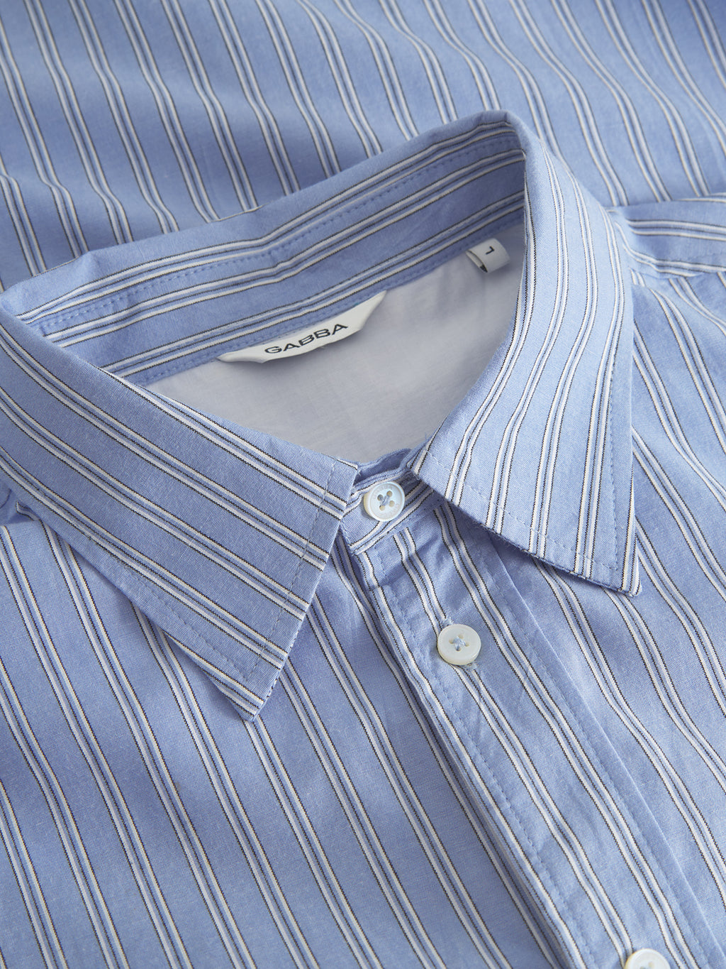 A close-up of the Gabba Sven Reg. Form LS shirt reveals a premium look with blue-striped button-up design, white buttons, and folded collar. Made from 100% cotton, it features thin vertical white and blue stripes and a visible white tag inside the collar for modern appeal.