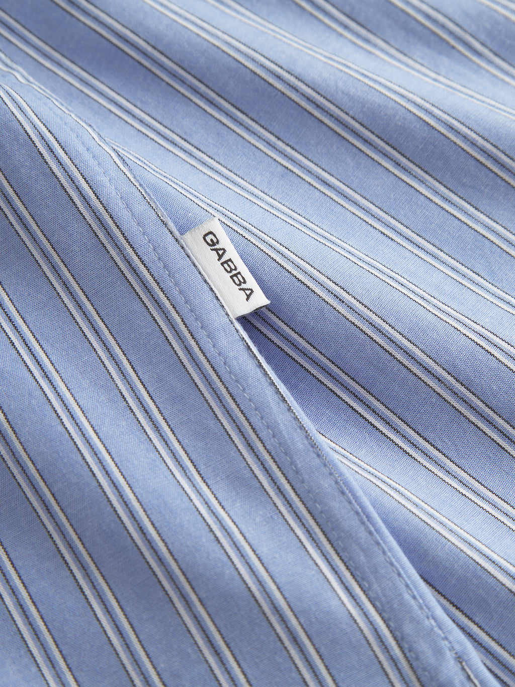 Close-up of a light blue fabric with thin white and darker blue stripes, ideal for a modern look. A label stitched diagonally across this 100% cotton shirt reads Diabea. Product: Sven Reg. Form LS - Blue Stripe by Gabba.