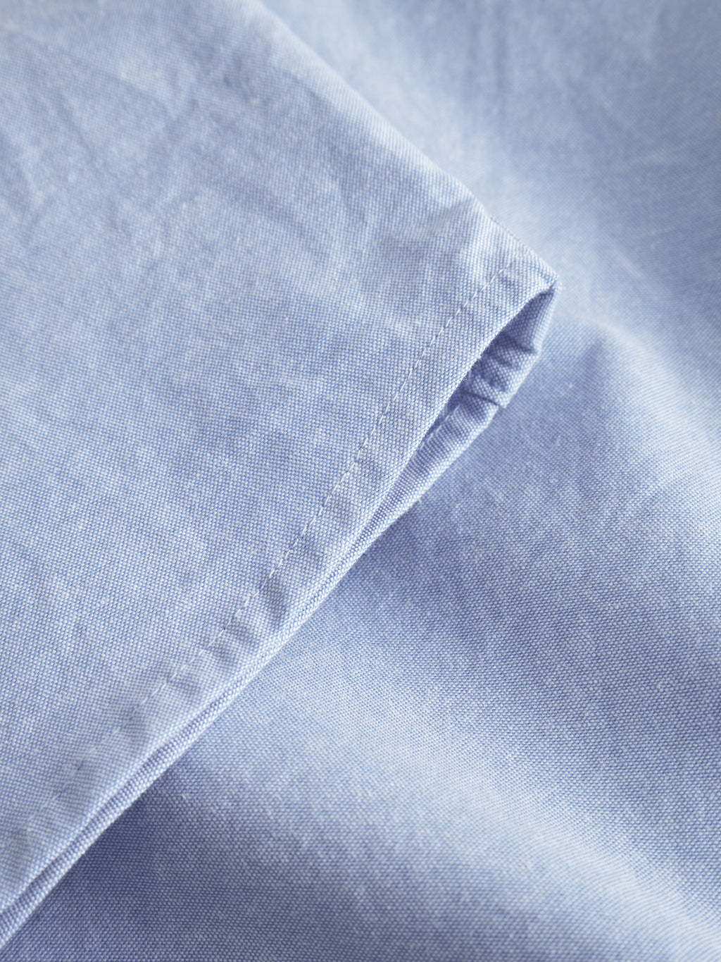 Close-up of light blue fabric with visible edge stitching, resembling the Bent Reg Ox LS - Light Blue by Gabba. The slightly wrinkled cotton creates a textured look offering all-day comfort.