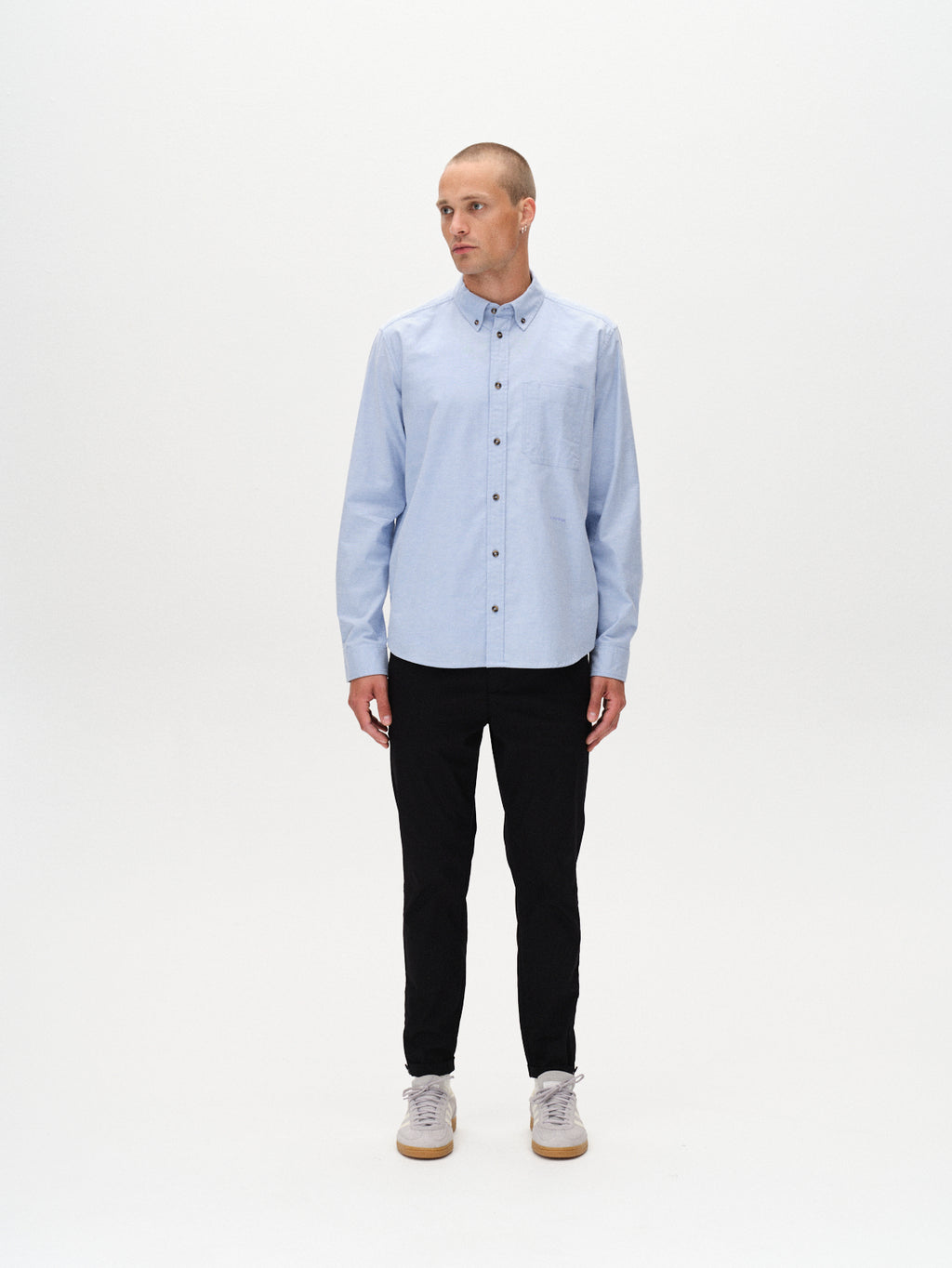 A man stands against a plain white background, wearing the Gabba Bent Reg Ox LS shirt in light blue with black pants and light gray sneakers. He faces slightly left, hands relaxed at his sides.