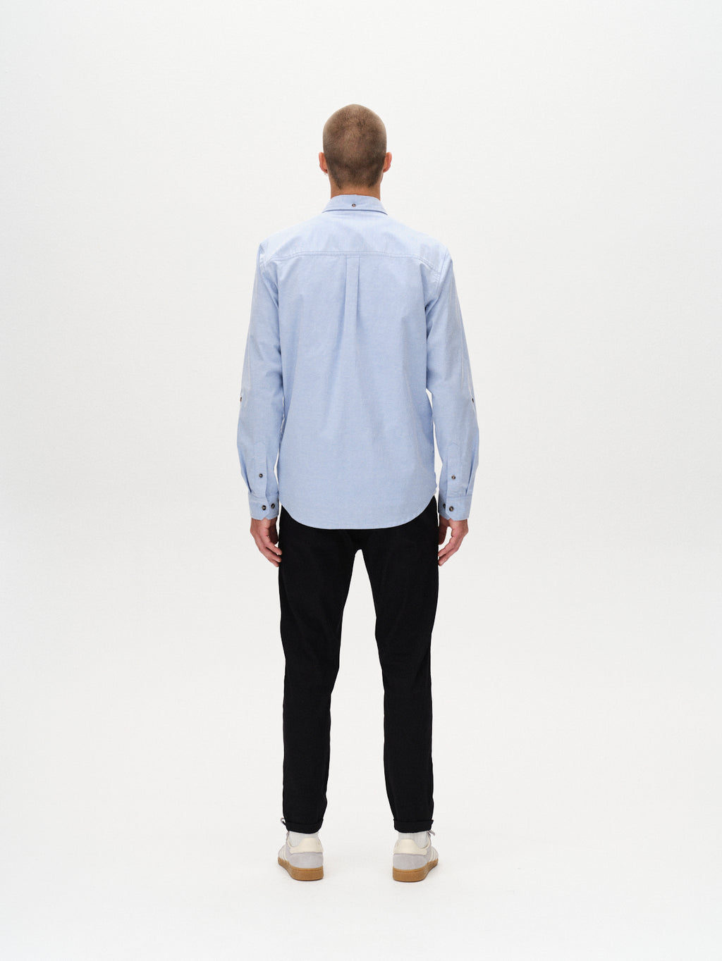 A man with short hair faces away from the camera, wearing the Gabba Bent Reg Ox LS shirt in light blue, black pants, and white shoes against a plain white background, highlighting the shirt’s versatile appeal.