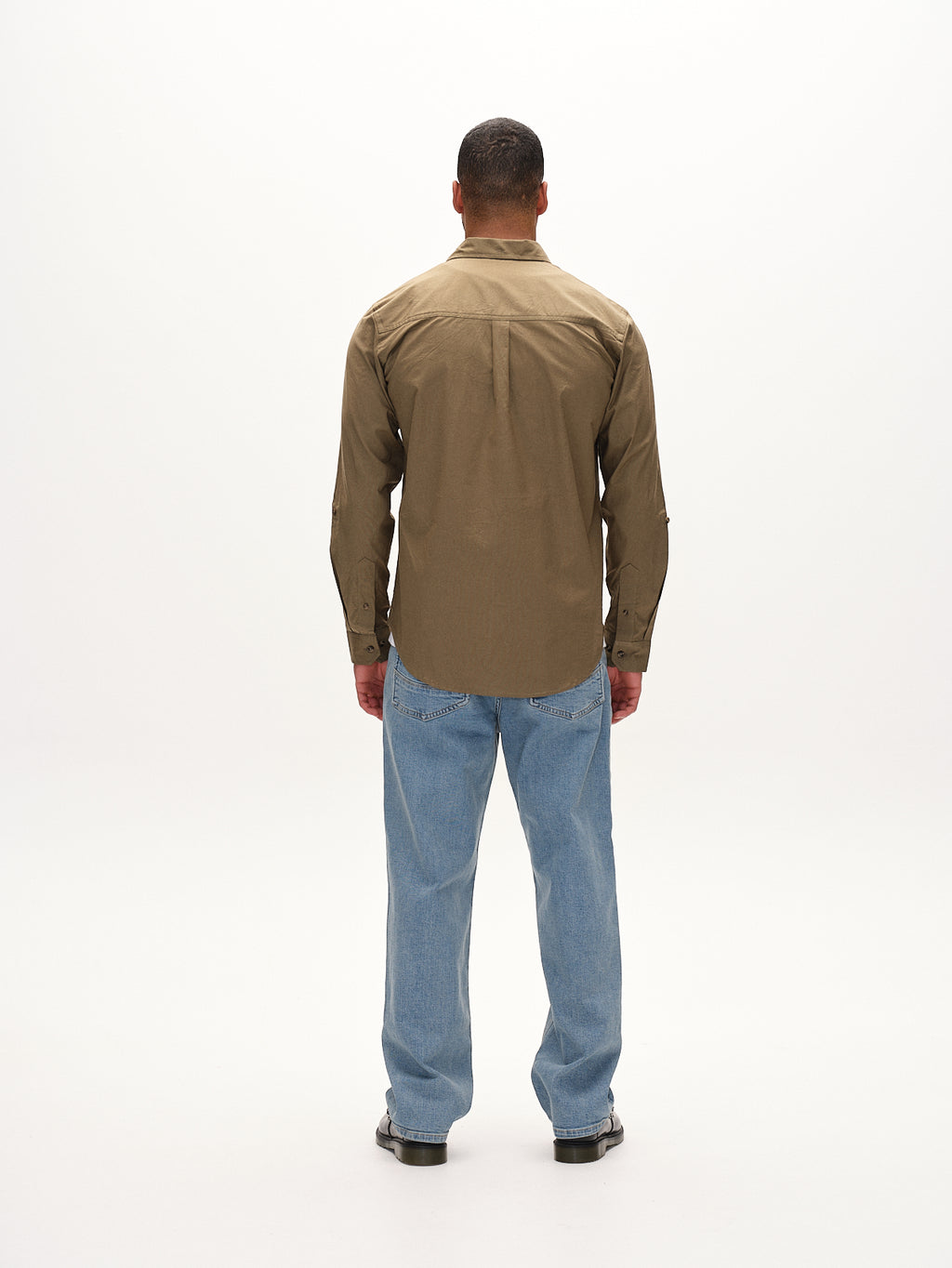 A man faces away, highlighting the Bent Reg Ox LS in army green by Gabba—a khaki button-up shirt with light blue jeans. His outfits timeless design and comfort blend seamlessly against the plain white background.