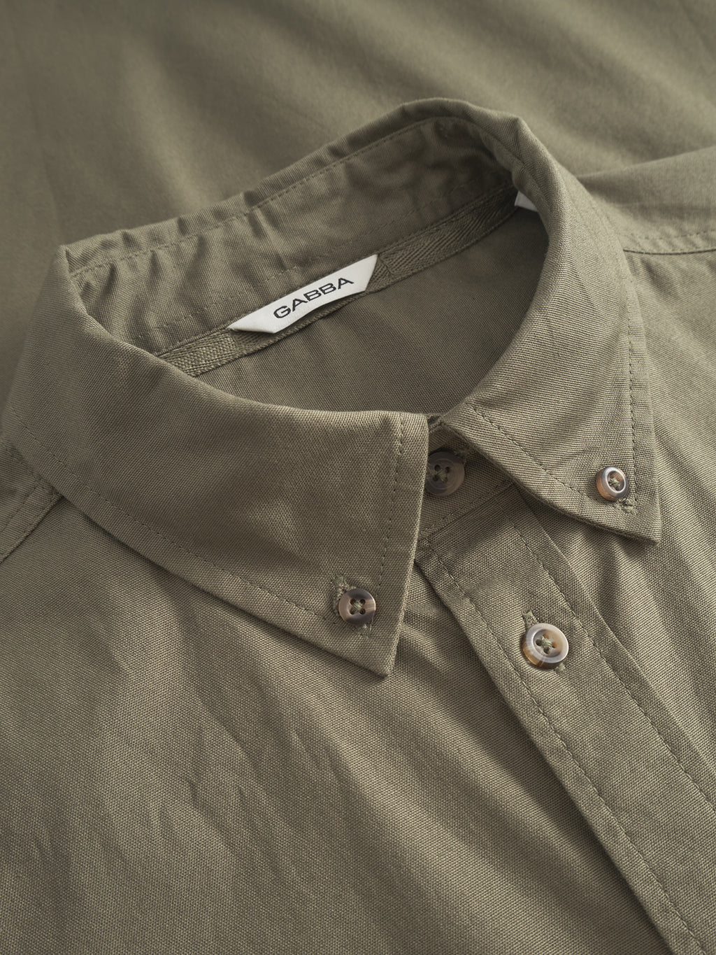 A close-up highlights a neatly folded Gabba Bent Reg Ox LS in Army, with a visible tag. The classic collar and button details enhance the slightly textured fabric, offering timeless design and all-day comfort for any occasion.