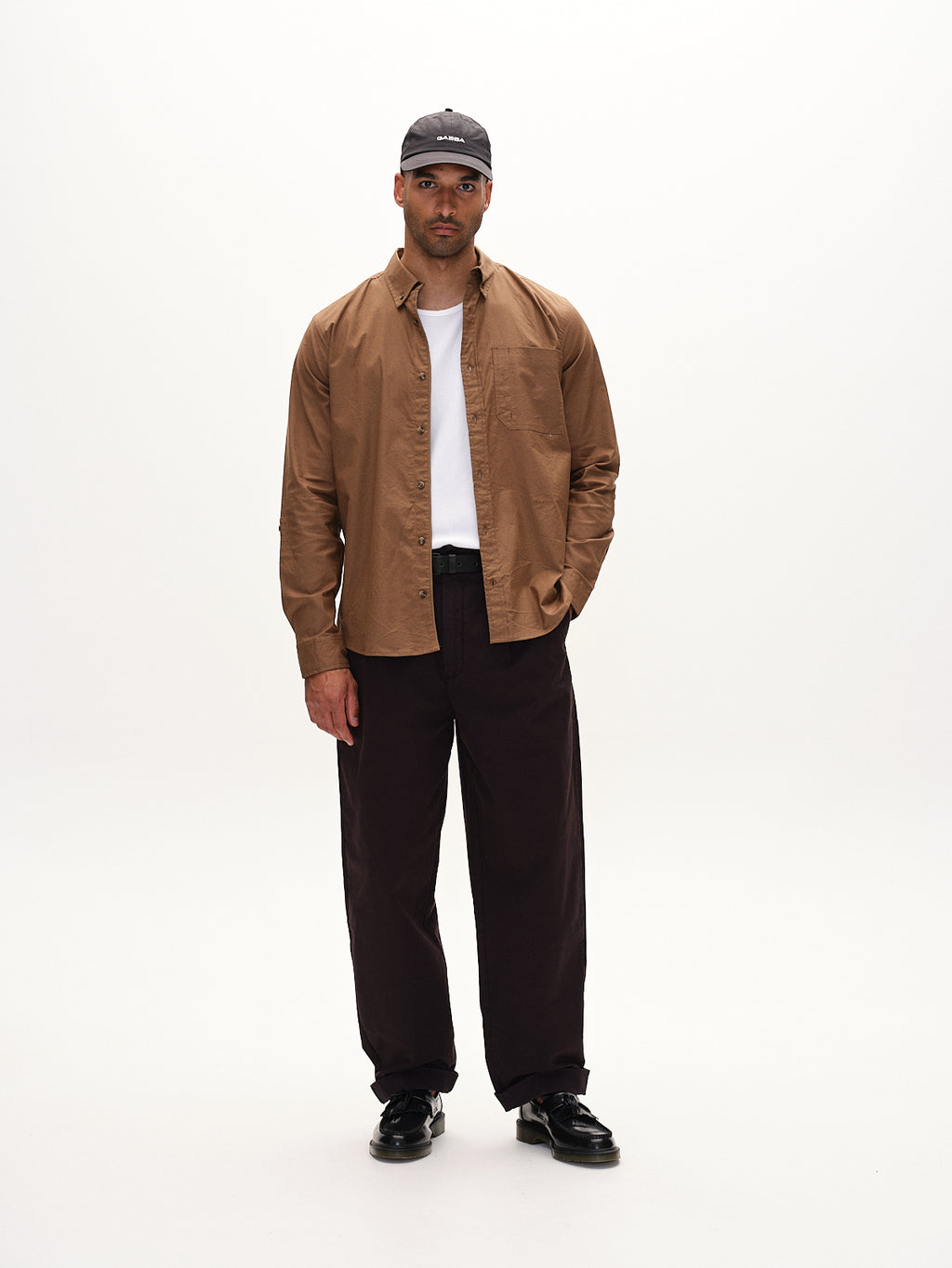A man stands against a plain white background wearing the Bent Reg Ox LS brown shirt by Gabba, a black cap, white T-shirt, black pants, and shoes. With one hand in his pocket and a neutral expression, this versatile design suits both formal and casual occasions seamlessly.