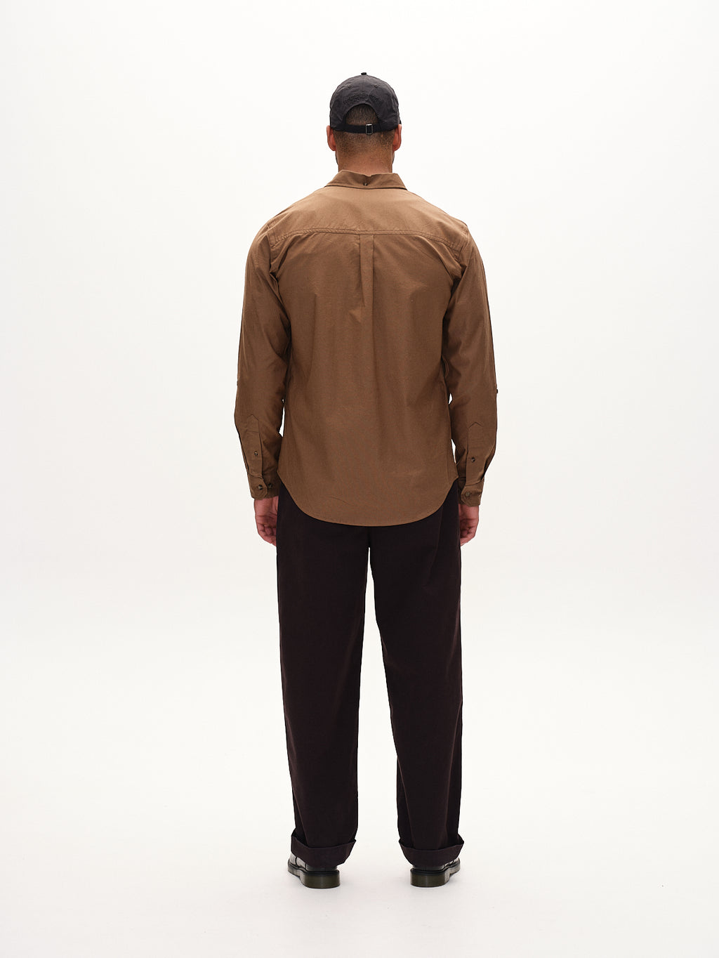 A person wearing Gabbas Bent Reg Ox LS in brown, paired with black pants and a black cap, stands facing away in a minimalistic, light-colored setting.