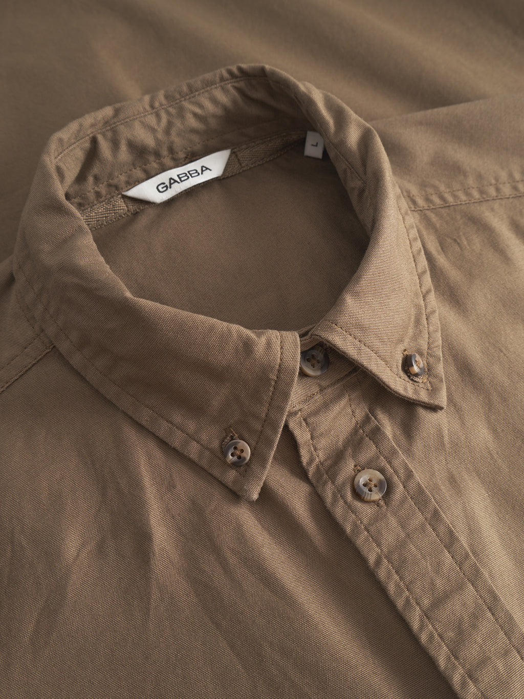 Close-up of a neatly folded Gabba Bent Reg Ox LS - Brown shirt, featuring a collared button-up cotton design. Displayed on matching fabric, it highlights its texture and stitching, ideal for formal and casual occasions.