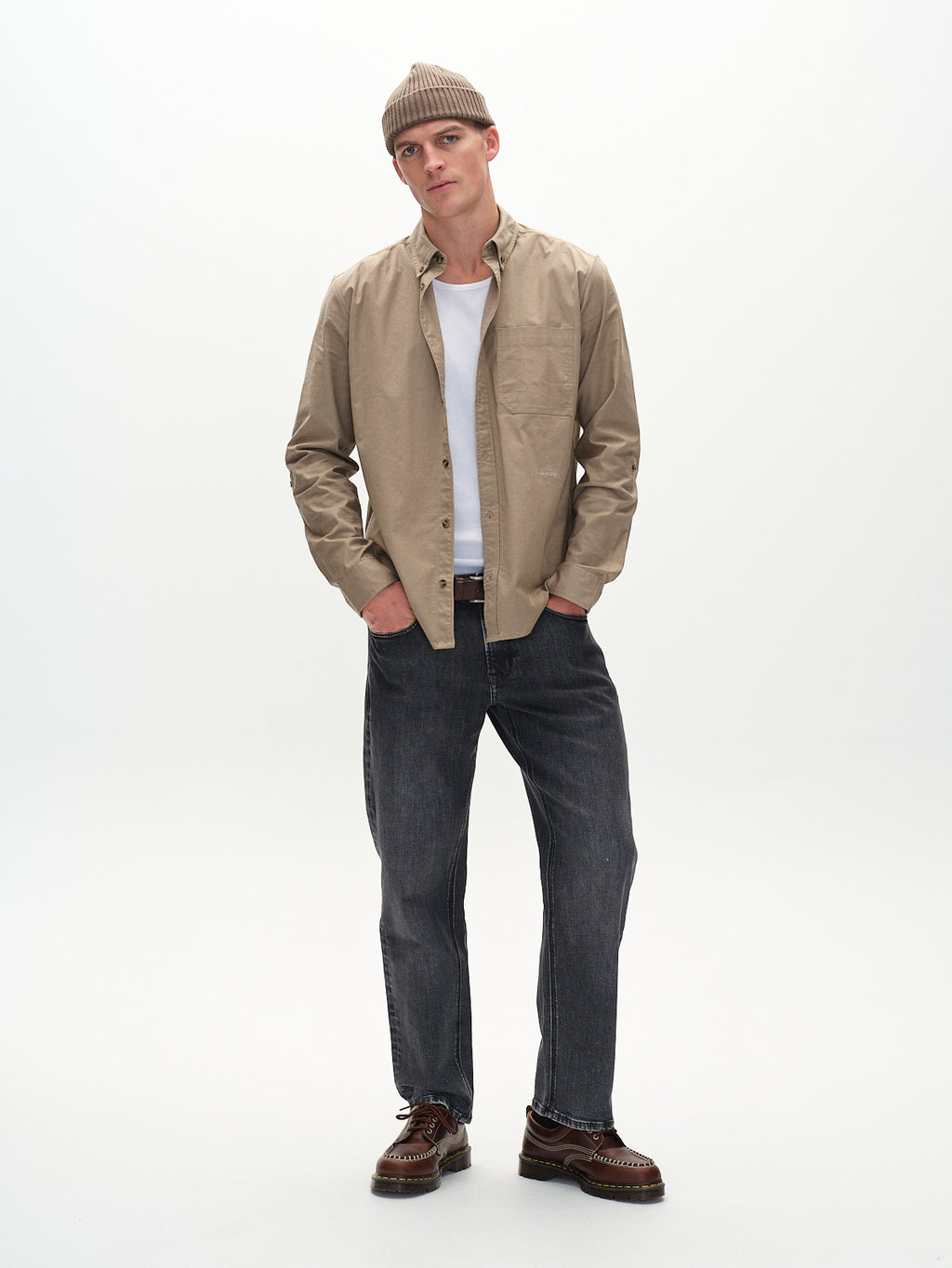 A man in a Gabba Bent Reg Ox LS Khaki shirt, beige beanie, white tank top, gray jeans, and brown shoes stands against a plain white background. His outfit combines classic elements for a timeless, versatile style.