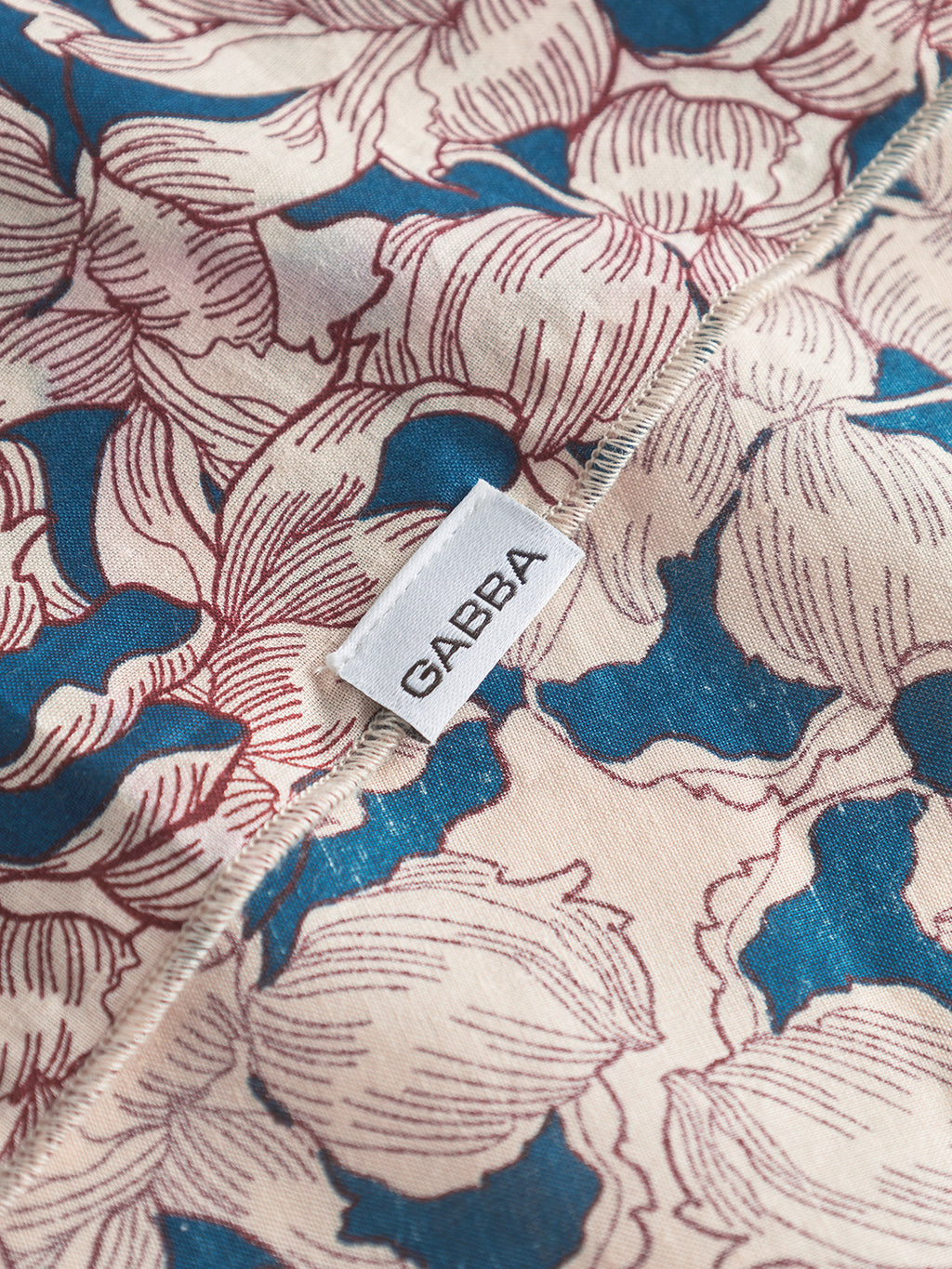 A close-up of the Bandana Epo - Blue Pattern by Gabba showcases a retro floral design in red and blue on beige. A white label with Gabba is stitched on the edge, emphasizing its elegant style.