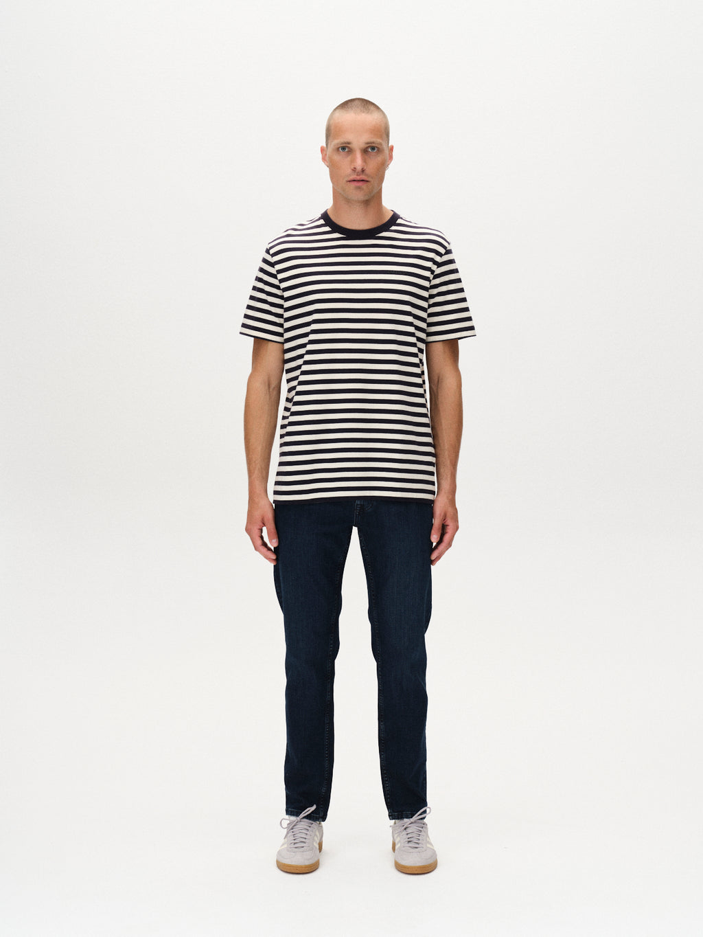 A man with a shaved head stands against a white background, wearing the Gabba Dune Logo Stripes SS GOTS - Navy Stripe t-shirt, dark blue jeans, and light gray sneakers. He faces forward with a neutral expression and relaxed posture.