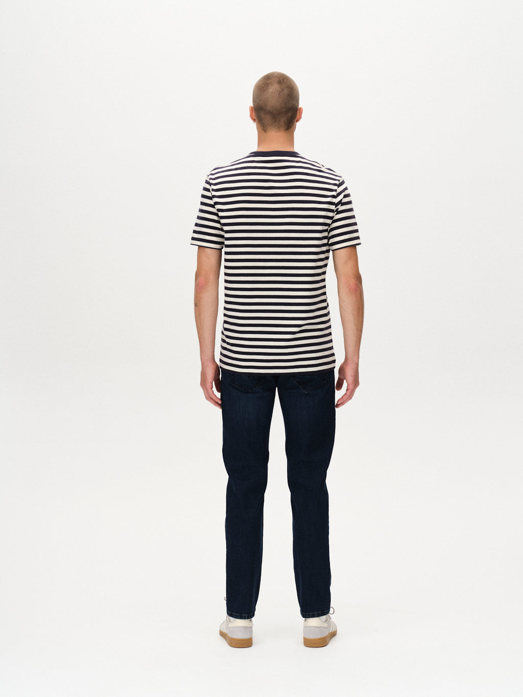 A person stands with their back to the camera in a Gabba Dune Logo Stripes SS GOTS - Navy Stripe shirt, paired with dark jeans and white sneakers, against a plain white background.