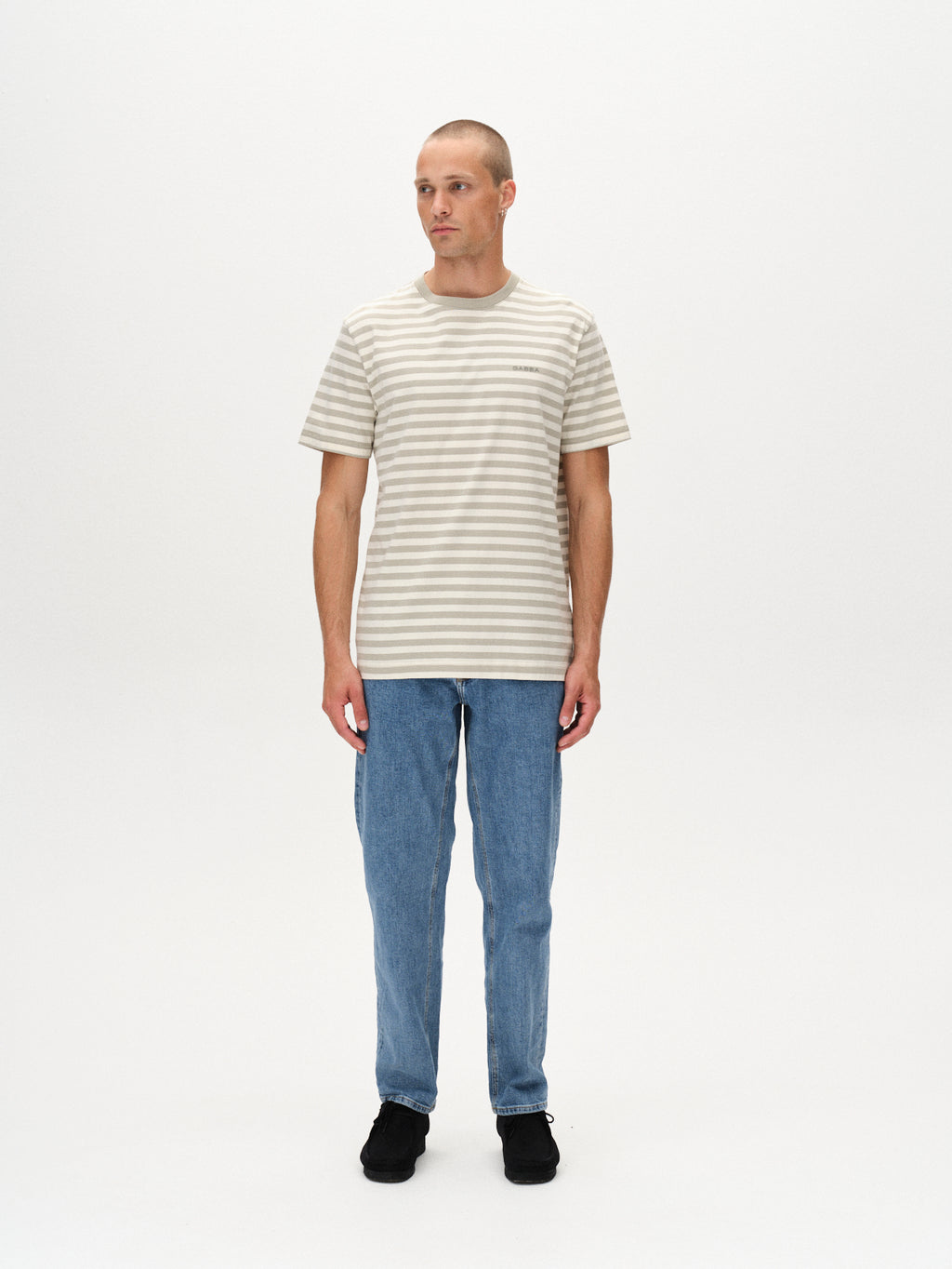 A man stands against a white background wearing the Gabba Dune Logo Stripes SS GOTS t-shirt in green stripe, paired with light blue jeans and black shoes. He looks slightly left with his arms relaxed at his sides.