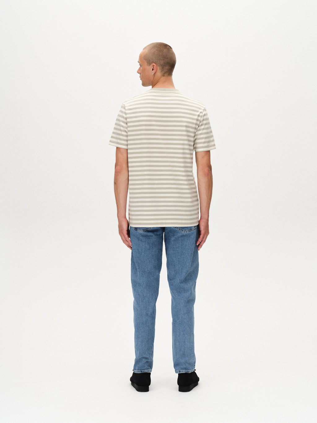 A person stands facing away from the camera, wearing the Gabba Dune Logo Stripes SS GOTS t-shirt in green stripes, blue jeans, and black shoes against a plain white background.