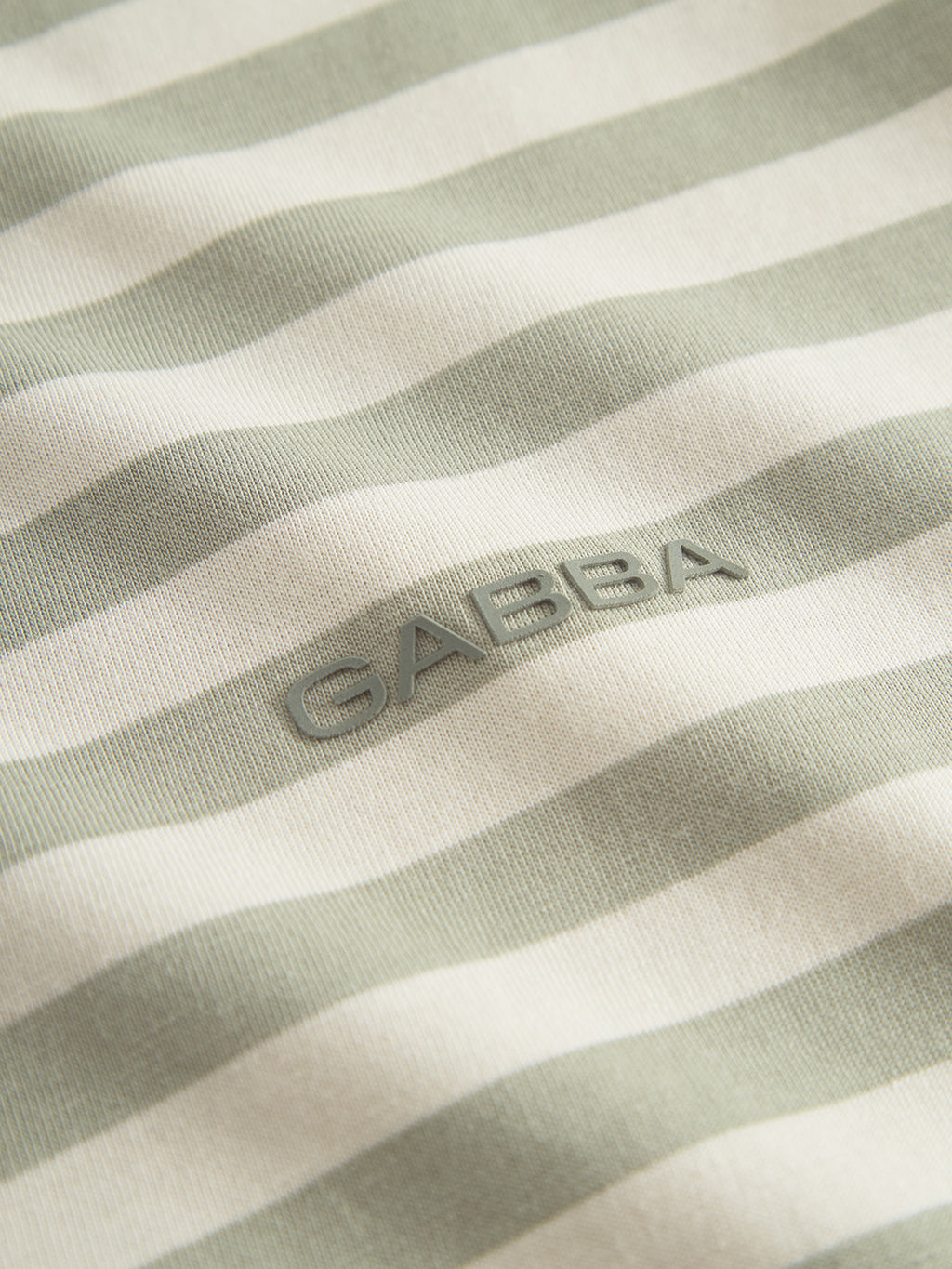 Close-up of the Dune Logo Stripes SS GOTS - Green Stripe fabric from Gabba, featuring horizontal light green and off-white stripes with raised GABBA lettering. This organic cotton, GOTS-certified fabric is perfect for crafting a stylish and sustainable basic t-shirt.