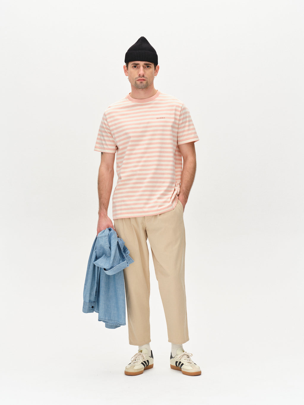 A man poses against a plain backdrop in the Gabba Dune Logo Stripes SS GOTS in Rose Stripes, featuring GOTS-certified organic cotton. His ensemble includes beige pants, a black beanie, a denim jacket in hand, and white sneakers with black stripes.