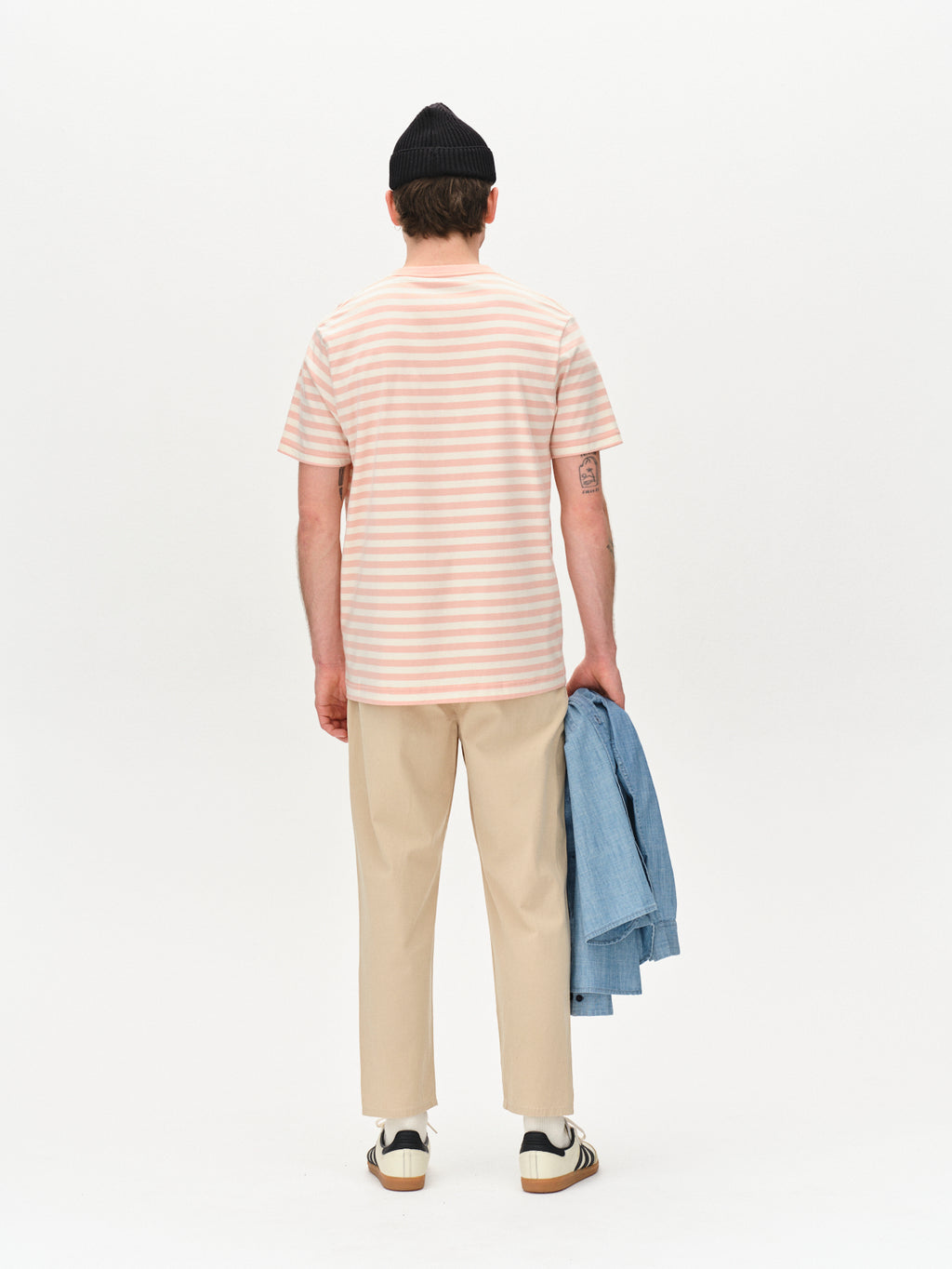 A person wearing a black beanie, beige pants, and sneakers holds a light denim jacket while facing away. Theyre styled in Gabba’s eco-friendly Dune Logo Stripes SS GOTS T-shirt in rose stripes. The plain white background highlights their fashionable yet sustainable look.