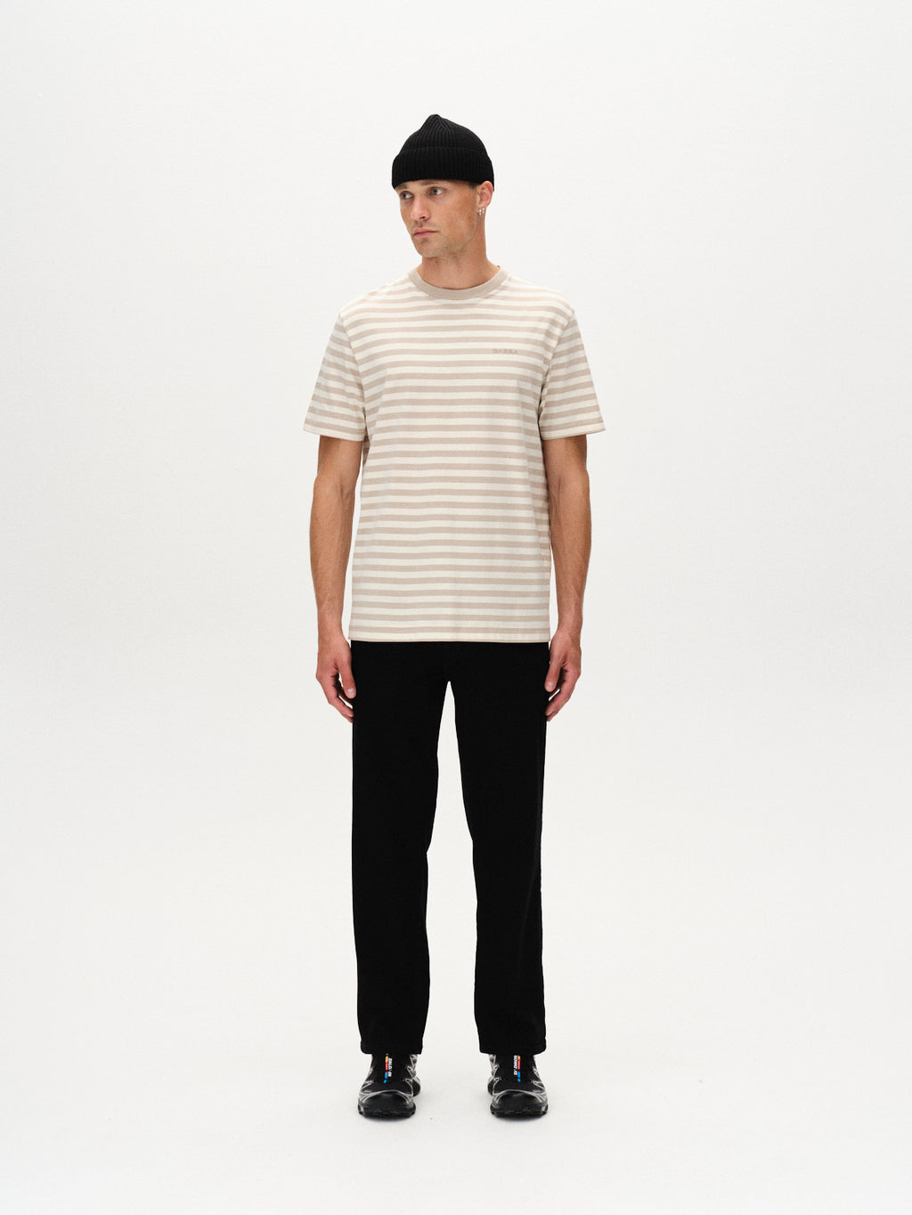 A person stands facing forward, wearing the Gabba Dune Logo Stripes SS GOTS t-shirt in beige stripe, paired with black pants, black shoes, and a black knit beanie. The background is plain white.