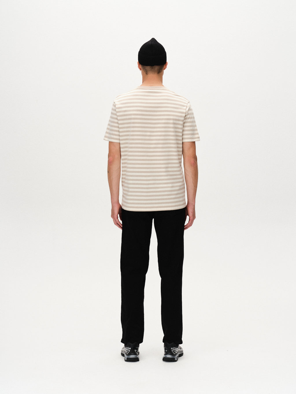 A person stands with their back to the camera, wearing Gabbas Dune Logo Stripes SS GOTS t-shirt in beige stripe, paired with black pants, shoes, and a beanie, against a plain white background.