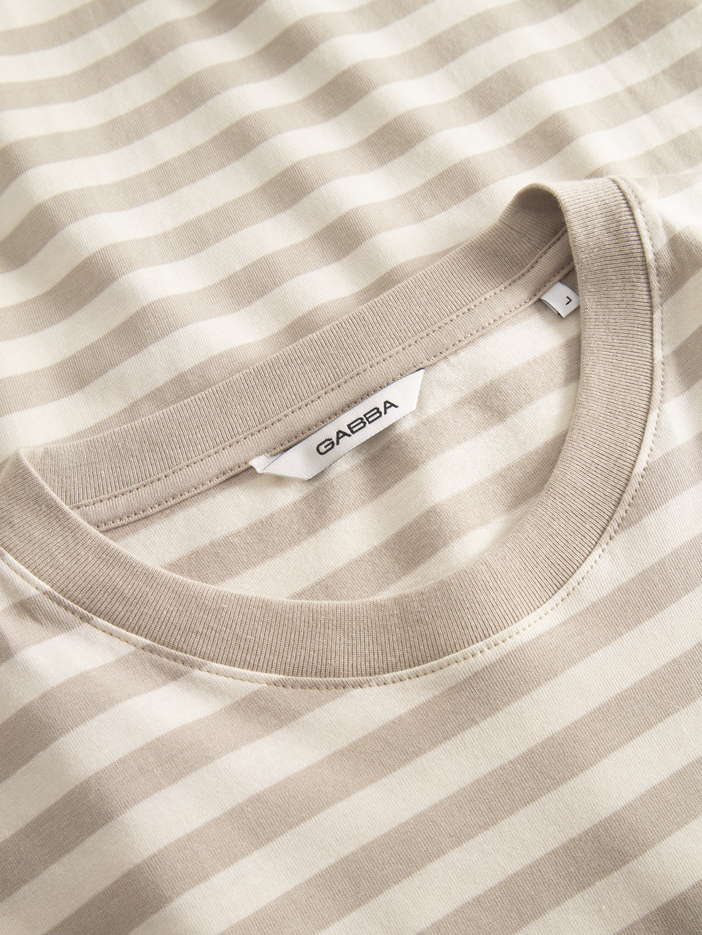 A close-up of the Dune Logo Stripes SS GOTS - Beige Stripe from Gabba reveals a classic fit, beige and white striped shirt with a round neckline, crafted from soft organic cotton. The label features Gabba in black on a white tag near the neckline, combining comfort and style.