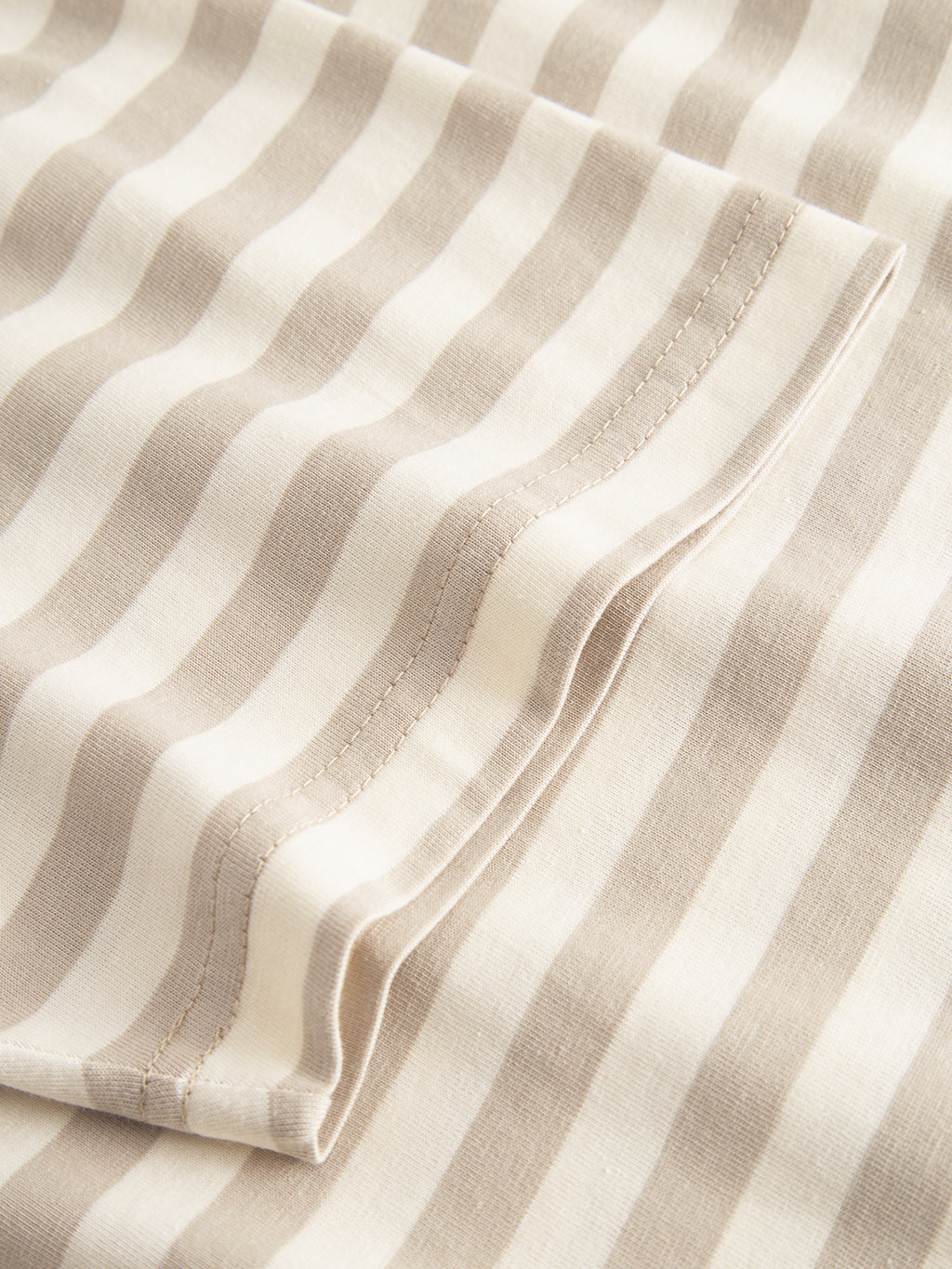 Close-up of Gabbas Dune Logo Stripes SS GOTS in beige stripe, a folded organic cotton fabric with beige and white stripes. It showcases noticeable edge stitching and a smooth, slightly wavy texture, ideal for a classic fit t-shirt.