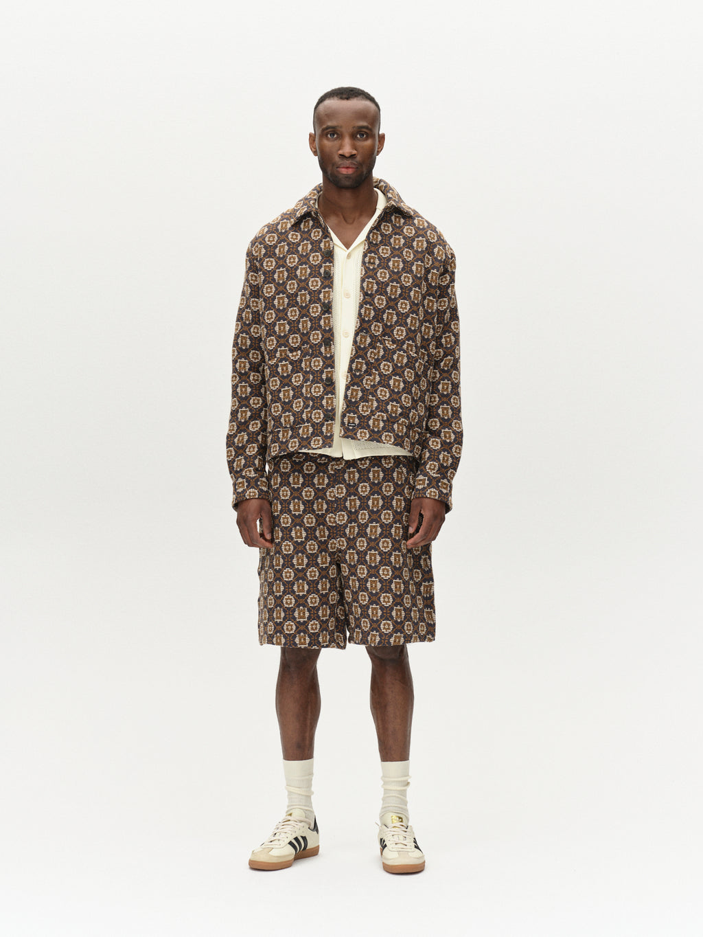 A person poses in the Fede Box Star ensemble by Gabba, featuring a geometric patterned jacket and shorts. The outfit is complemented by a white shirt and sneakers with black stripes, highlighting a brown and beige color scheme against a plain backdrop.