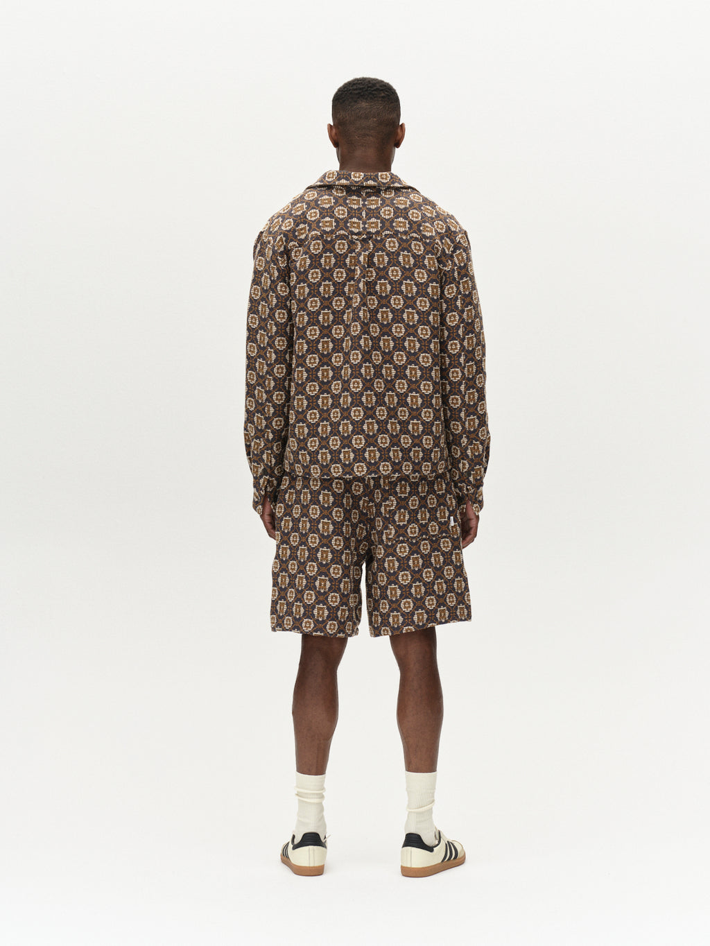 A person stands with their back to the camera wearing Gabbas Fede Box Star, a striking multi-patterned jacket and boxy shorts in brown and beige tones. Their short hair accents white socks and beige and black sneakers against a plain white background, highlighting the ensembles bold statement.