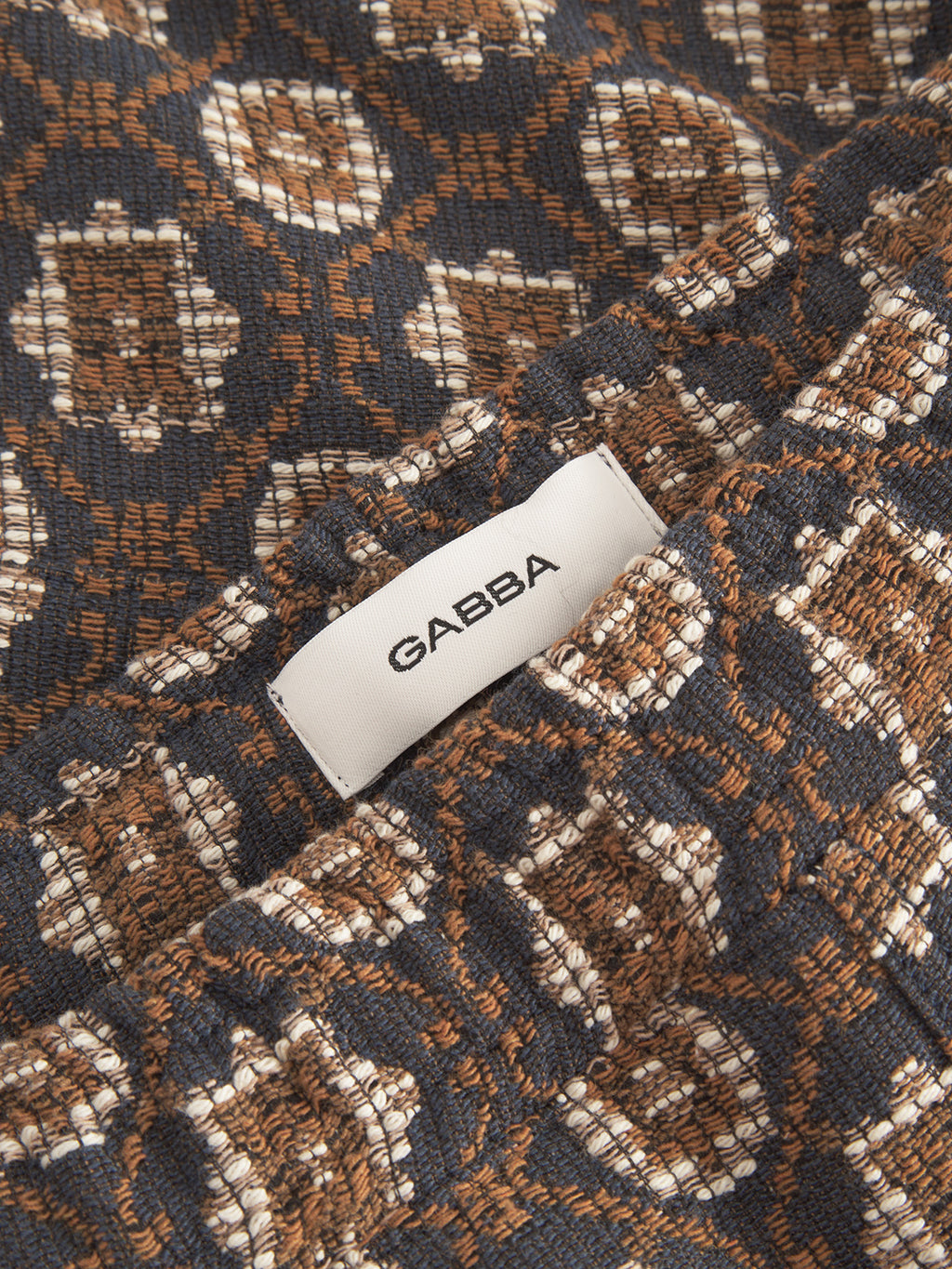 The Fede Box Star - Multi Pattern from Gabba features a textured fabric with a geometric pattern in brown, navy, and cream. A small white Gabba label is sewn on, partly hidden by folds. This design enhances any statement outfit or loose fashion ensemble.