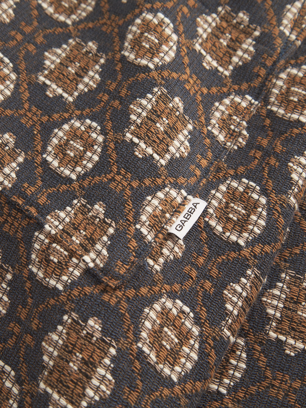 Close-up of a textured fabric from Fede Box Star - Multi Pattern by Gabba, featuring a brown and cream geometric design on a dark background. A small white label with Gabba is sewn onto the fabric, perfect for elevating any statement outfit.