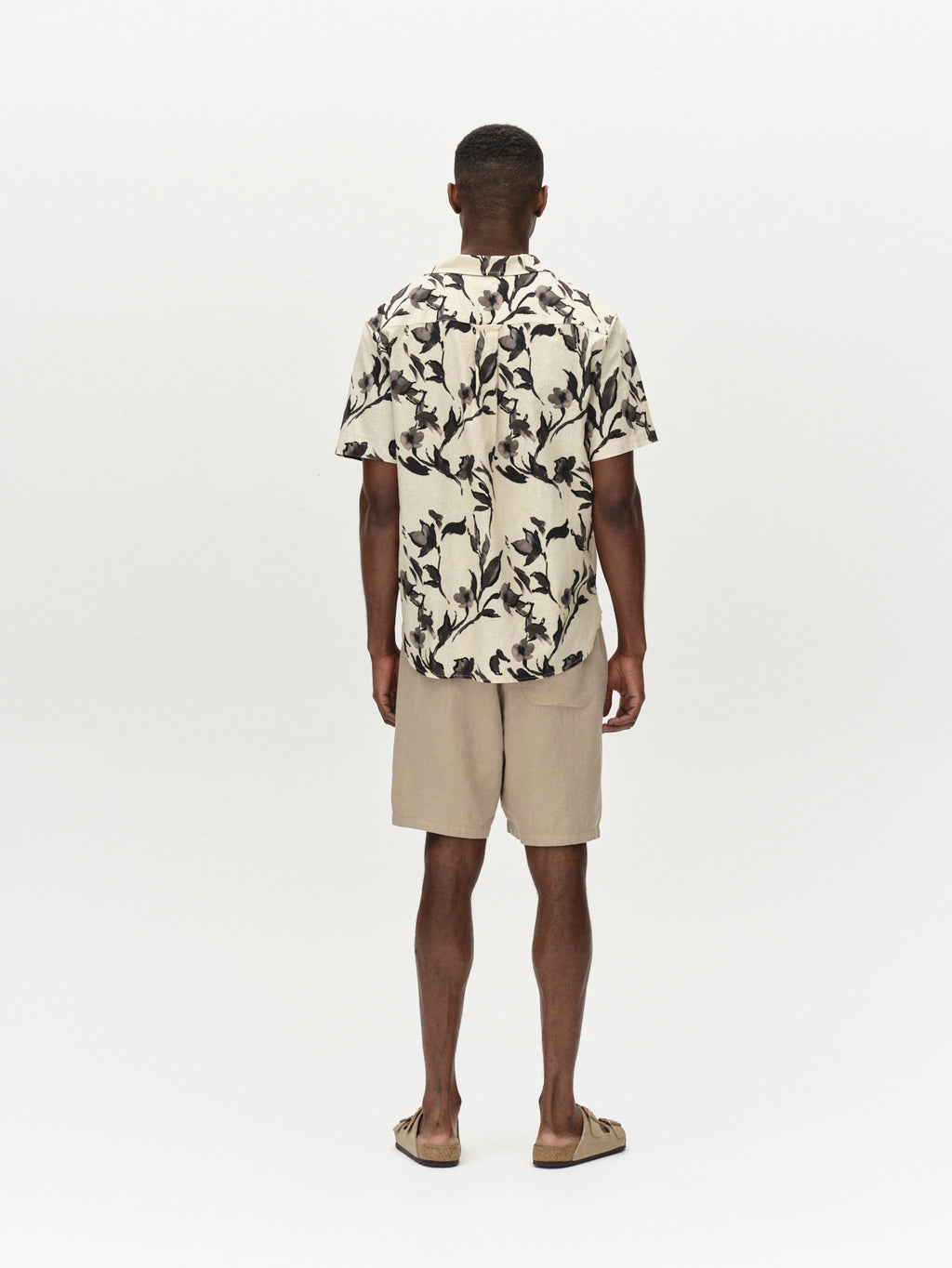 A man faces away from the camera wearing the Gabba Sven Reg Resort Bloom SS - Meteorite shirt, beige shorts, and brown sandals against a plain white background.