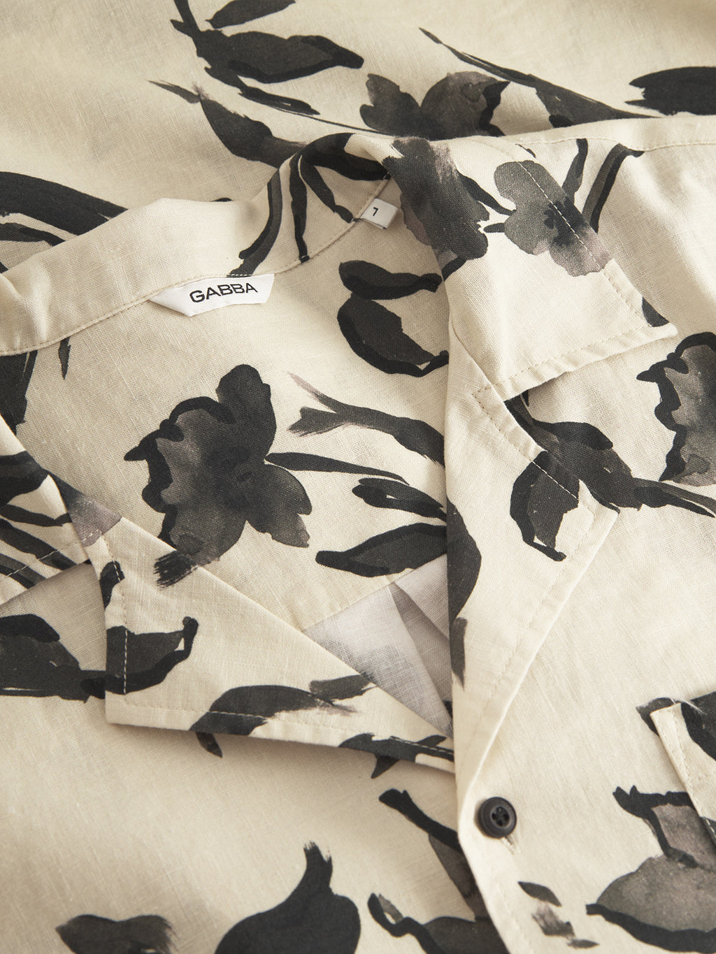 A close-up of the Sven Reg Resort Bloom SS shirt by Gabba shows a beige premium design with a black flower print. It features a collar, partially unbuttoned, displaying the GABBA label inside. Made from quality linen, its smooth and lightweight.