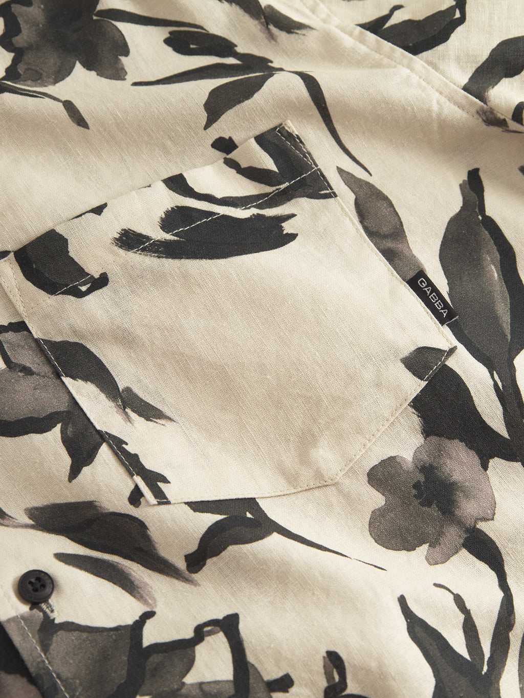 Close-up of the Sven Reg Resort Bloom SS - Meteorite shirt by Gabba in beige, featuring a black and gray flower print. The high-quality linen fabric has a subtle sheen, with a left front pocket adorned with a small GAIEA tag and partially visible buttons below.