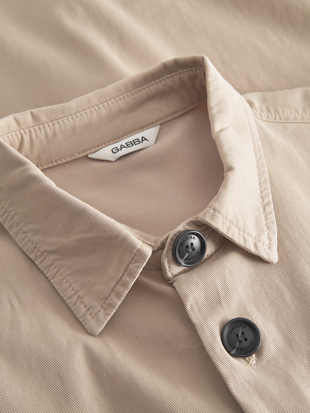 A close-up of the Gabba Jens Over Lubo LS in sand reveals button details and a Gabba label. The summer fabric has a subtle texture for comfort, with neatly arranged collar and buttons. The shirt is flat on a matching sand surface.