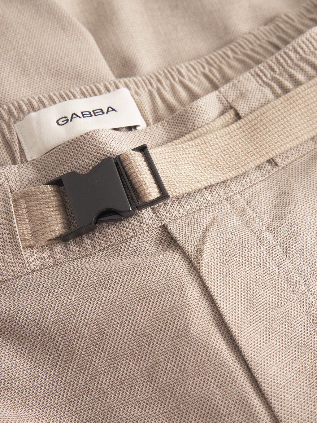 Close-up of Gabba’s Coz Belt Carre Shorts in sand, featuring superior comfort, an elastic waistband, a unique belt strap with plastic buckle, and a white Gabba label.