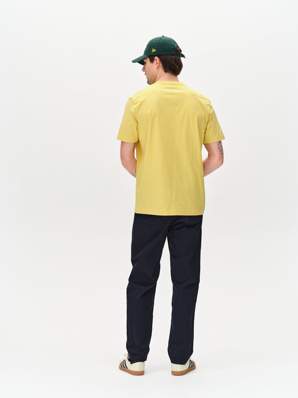 A person models the Joe Unite - Navy from Gabba, pairing a yellow t-shirt and dark tapered cotton pants. A green cap completes the look against a plain white backdrop, highlighting the casual style.