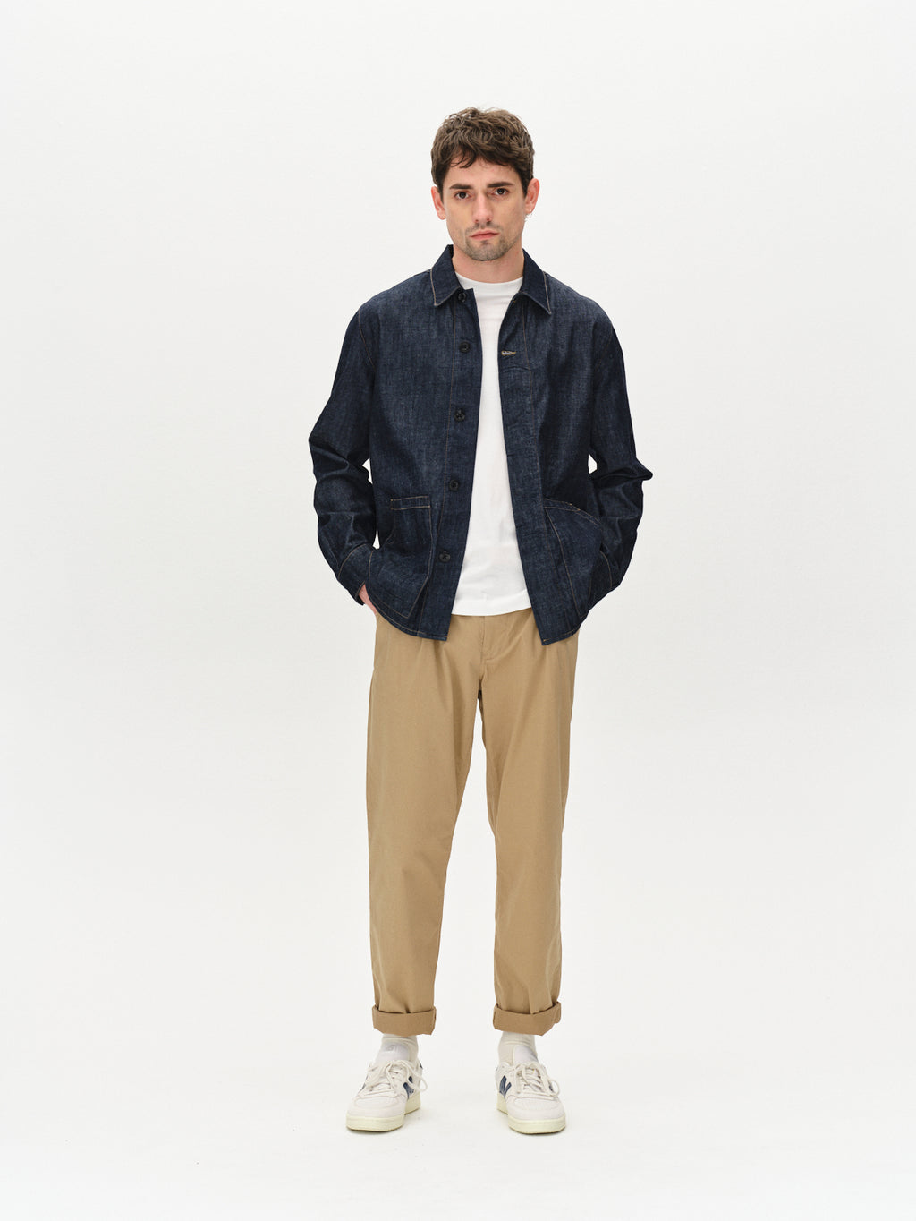 Against a plain backdrop, a man wears a dark denim jacket over a white shirt and relaxed-fit pants for a casual look. His ensemble is completed with Gabbas Joe Unite - Sand tan tapered pants and white sneakers; he gazes forward with hands in pockets, maintaining a neutral expression.