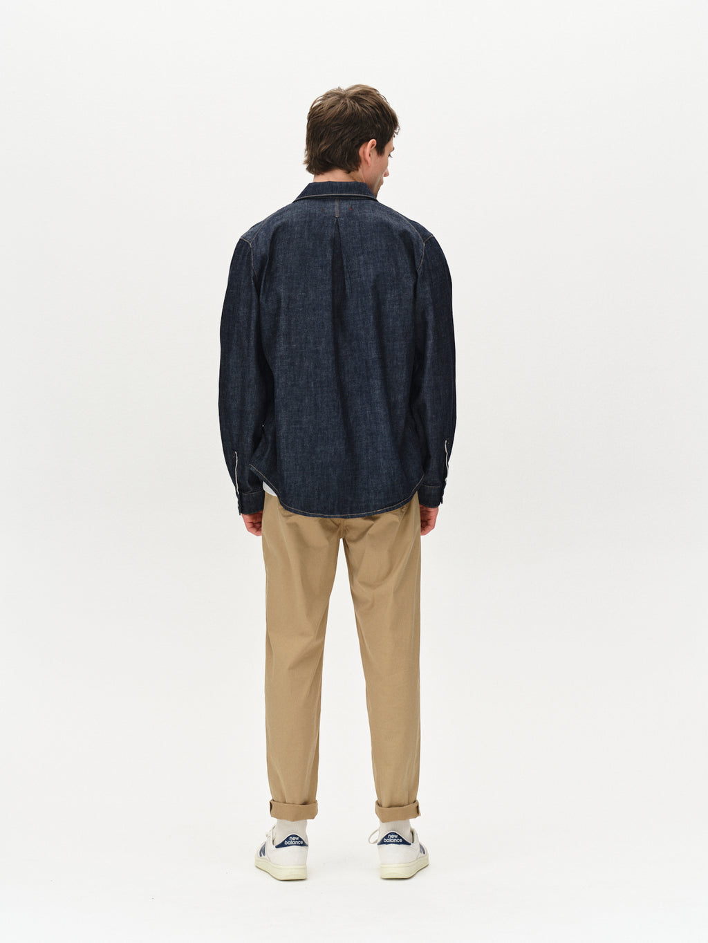 A person faces away from the camera, flaunting a casual vibe in a dark denim shirt, Gabbas Joe Unite - Sand tapered-leg pants, and white sneakers against a plain white backdrop, emphasizing the relaxed style.