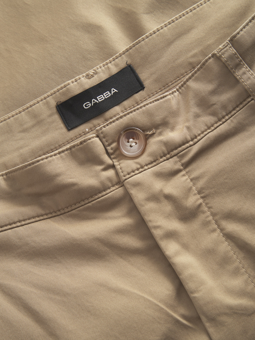Close-up of Joe Unite - Sand pants by Gabba featuring a relaxed fit, visible button, and stitching details. The smooth fabric complements a casual look, while the neatly tailored design with tapered legs adds modern appeal. A black Gabba label is attached inside the waistband.