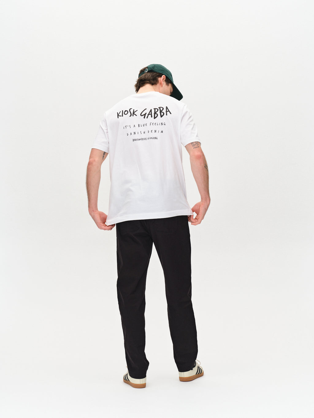 A person wears the Gabba white T-shirt with contrasting text, Joe Unite - Black relaxed pants, and a dark green baseball cap. The back of the T-shirt has additional text below the main logo, creating a casual look against a plain white backdrop.