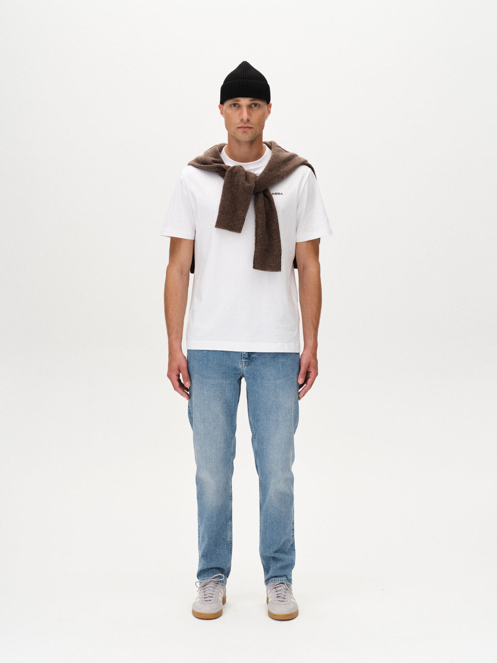 A man faces forward against a white background, wearing a black beanie, white T-shirt, Marc 5poc - Mid Blue Denim jeans by Gabba, white sneakers, and a brown sweater draped over his shoulders for a modern classic look.