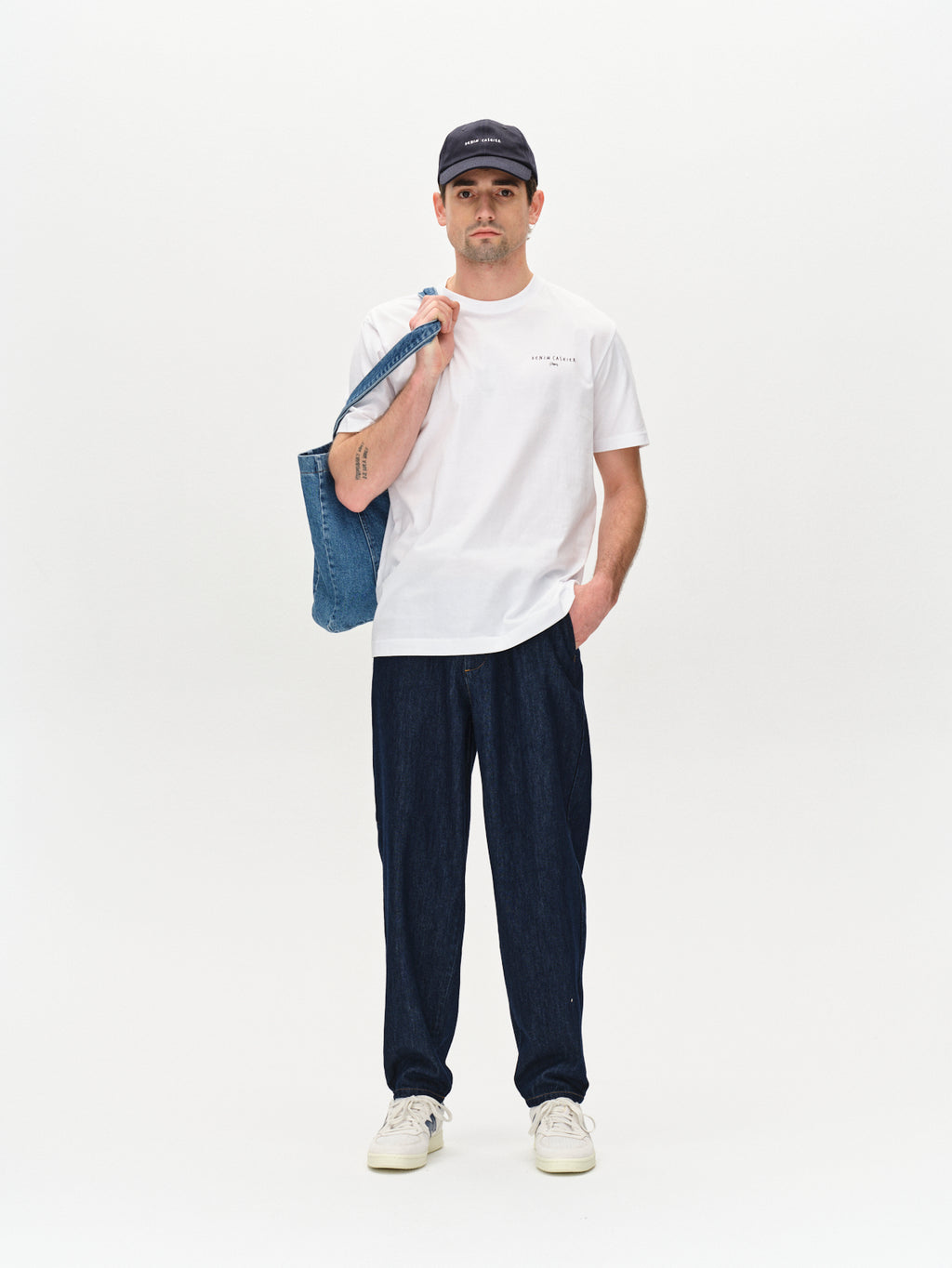 A person in a white T-shirt, Vito - Dark Blue Denim by Gabba jeans, and classic white sneakers finishes their look with a black cap. They stand against a plain backdrop, showcasing workwear-inspired style while effortlessly carrying a denim tote bag over one shoulder.