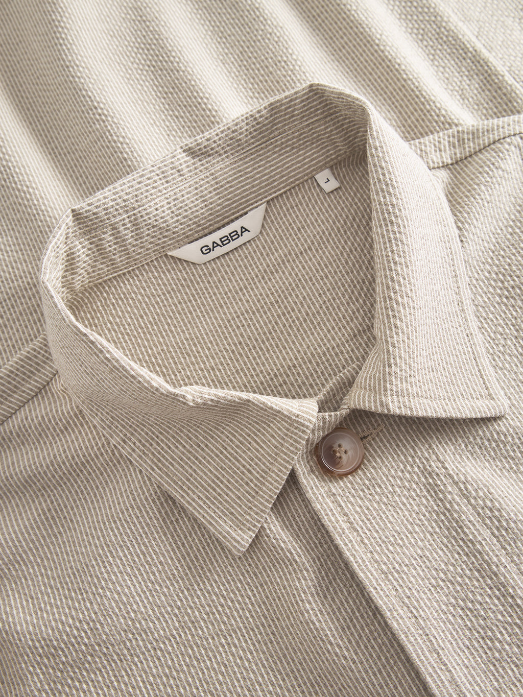 A close-up of the textured, light beige Gabba Today Com2 - Khaki button-up shirt shows a collar and fastened button. Its thin vertical stripes and seersucker fabric add elegance, hinting at thoughtful overshirt styling.