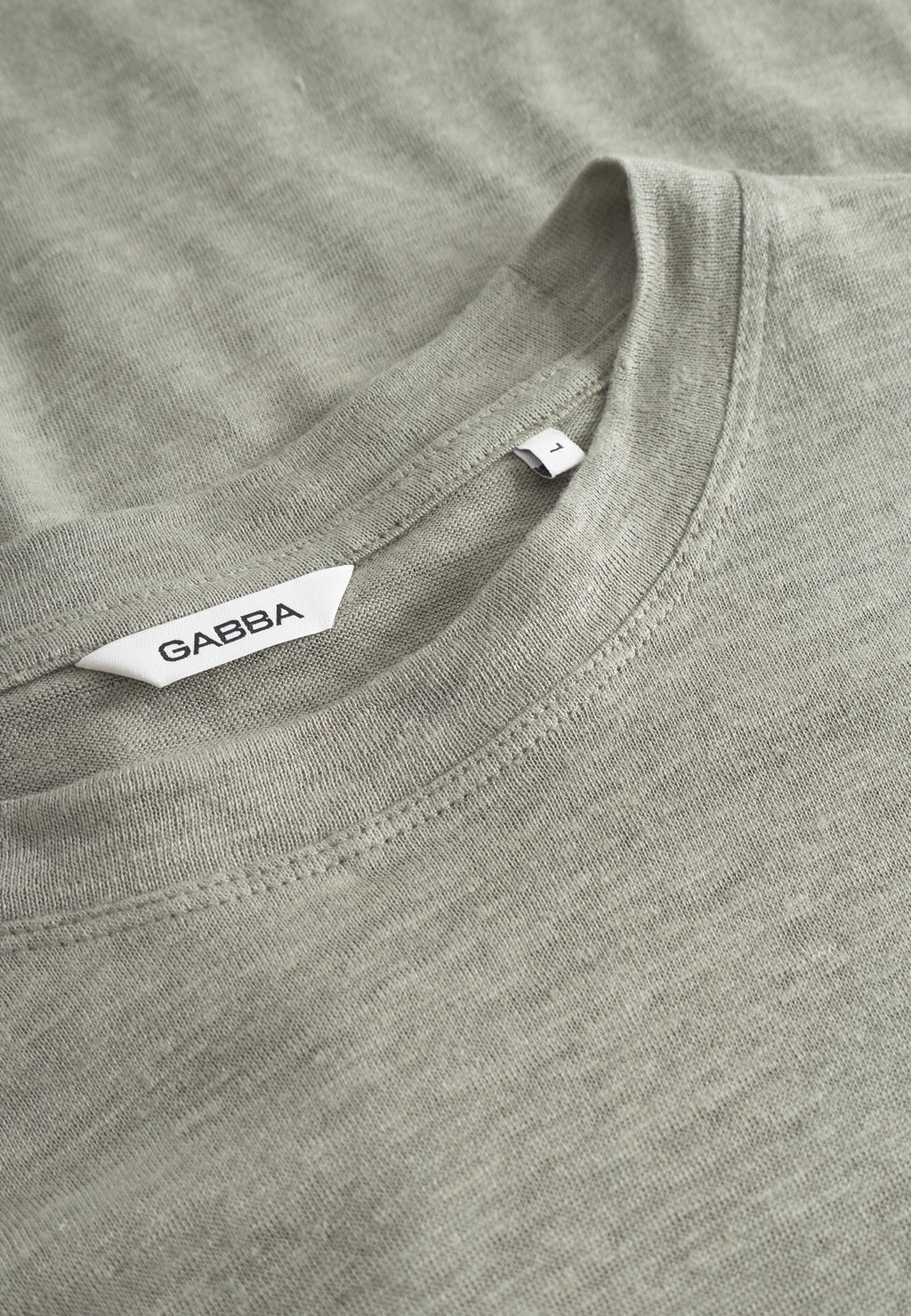 Close-up of a light green t-shirt lying flat, highlighting its round neckline and a tag labeled Gabba. The material looks soft with slight texture, featuring tone-on-tone stitching for subtle detail on this stylish Duke Linen Logo SS piece.