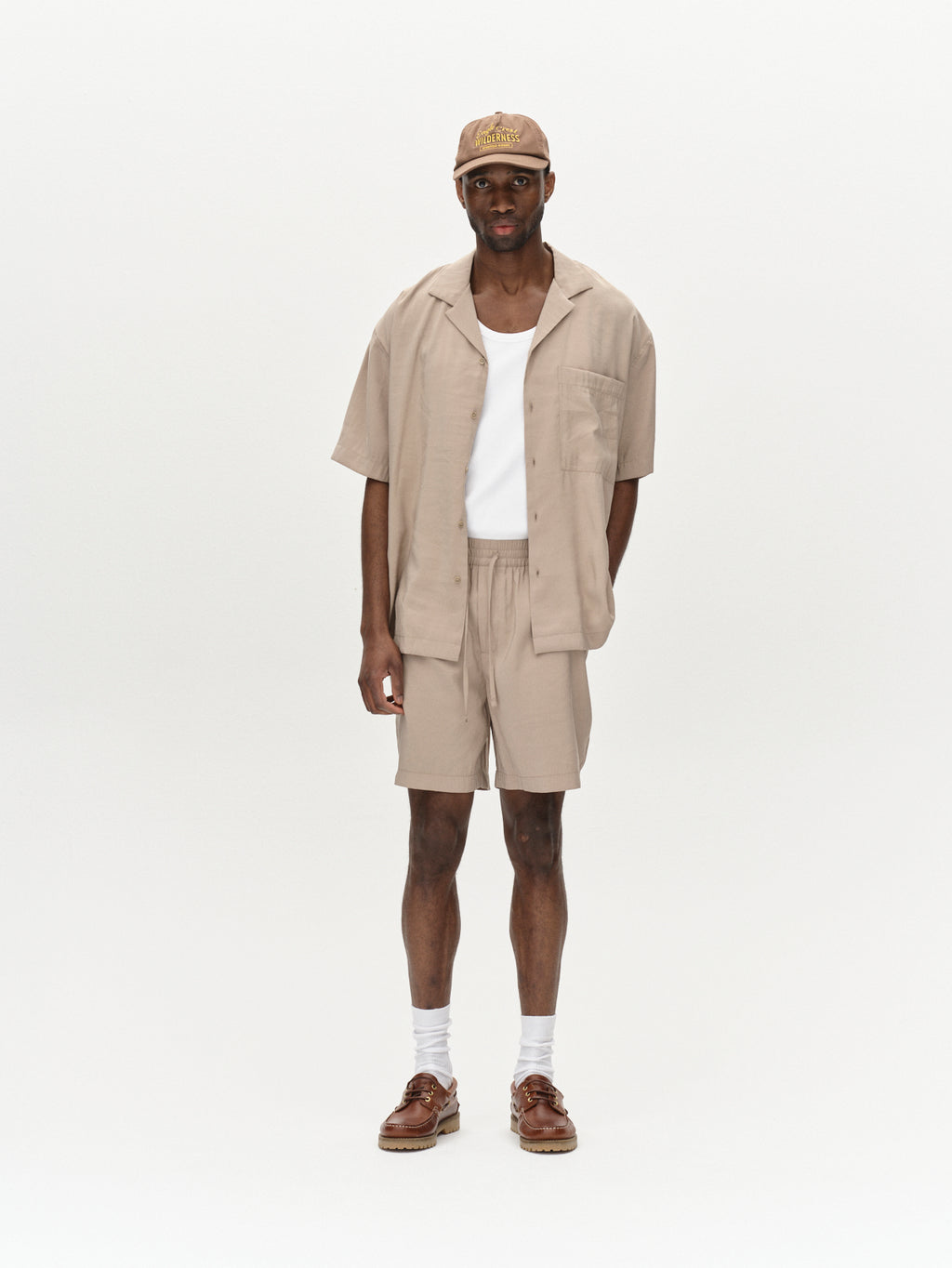 In front of a plain white backdrop, someone is dressed in the Bo Yari - Sand Greige light brown open short-sleeve shirt and matching premium shorts from Gabba. They complete the look with a cap and brown shoes paired with white socks for superior comfort on casual outings.