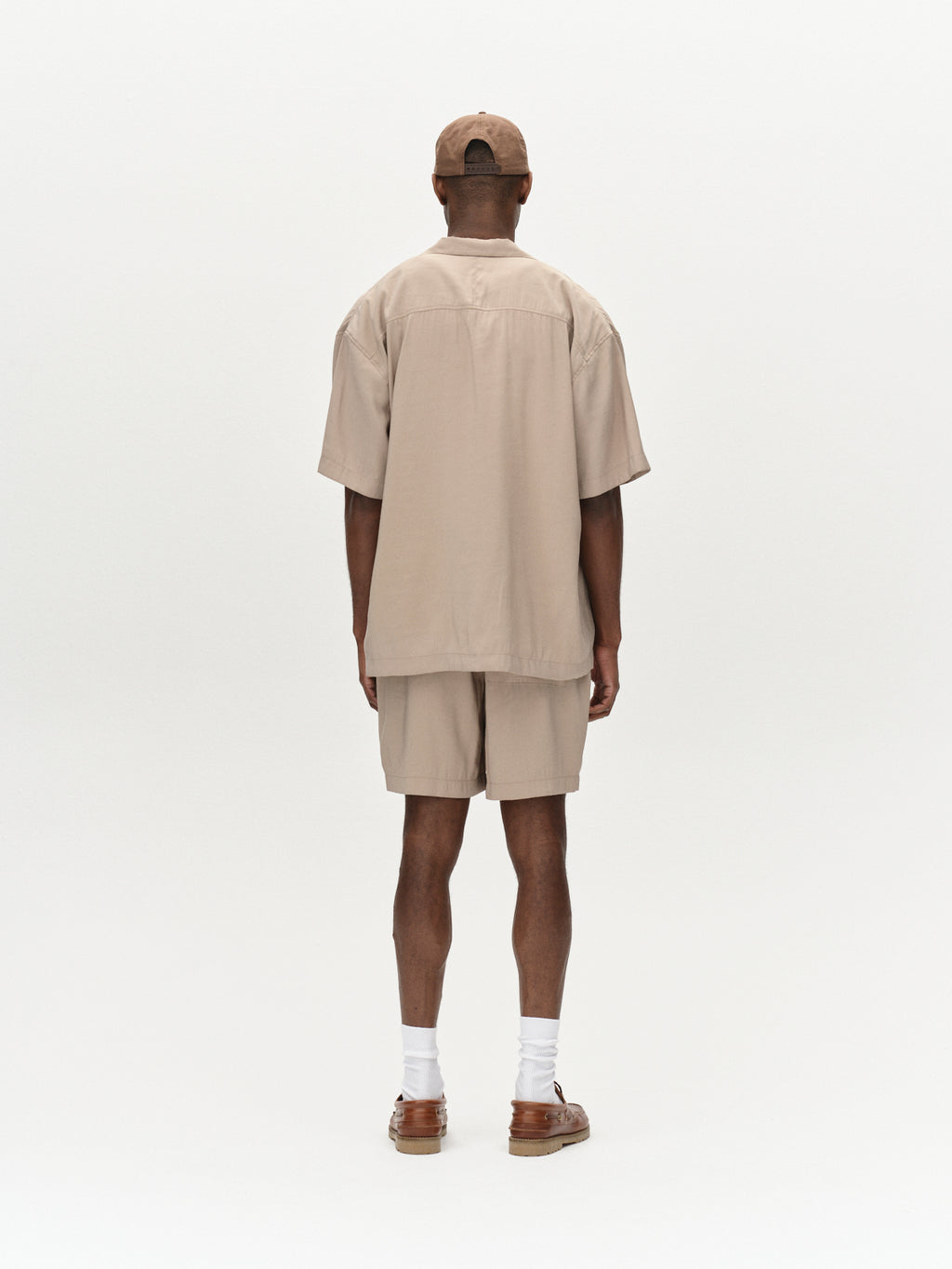 Facing away on a plain white background, a person wears Gabbas Bo Yari in sand greige—a beige shirt with premium lightweight shorts—paired with a cap, white socks, and brown sandals.