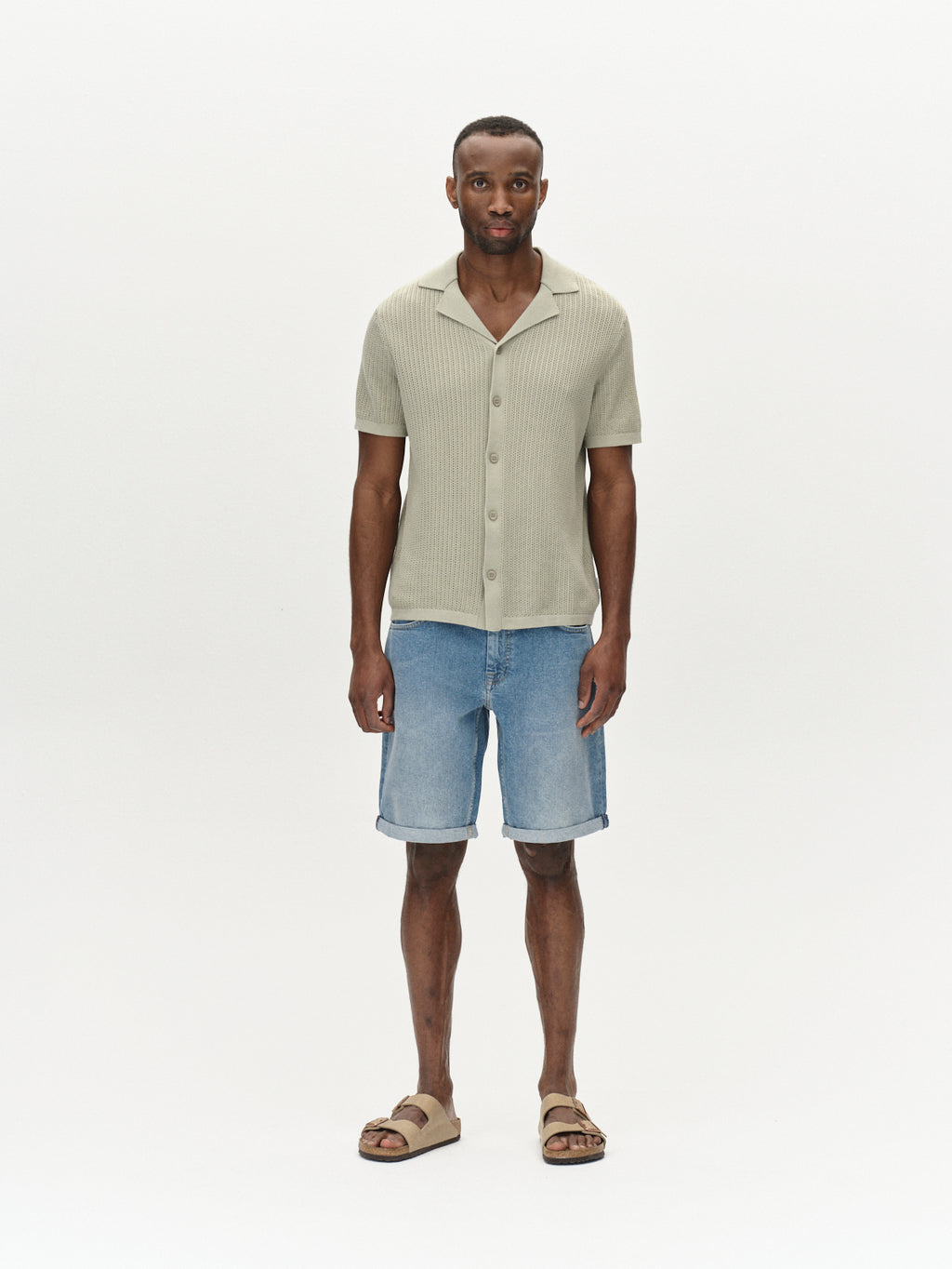 A man stands against a plain background wearing Gabbas Markus 5poc mid-blue denim shorts and a light green short-sleeve button-up shirt made from recycled viscose. His beige sandals add to the comfortable fit, while he maintains a neutral expression with his arms relaxed at his sides.