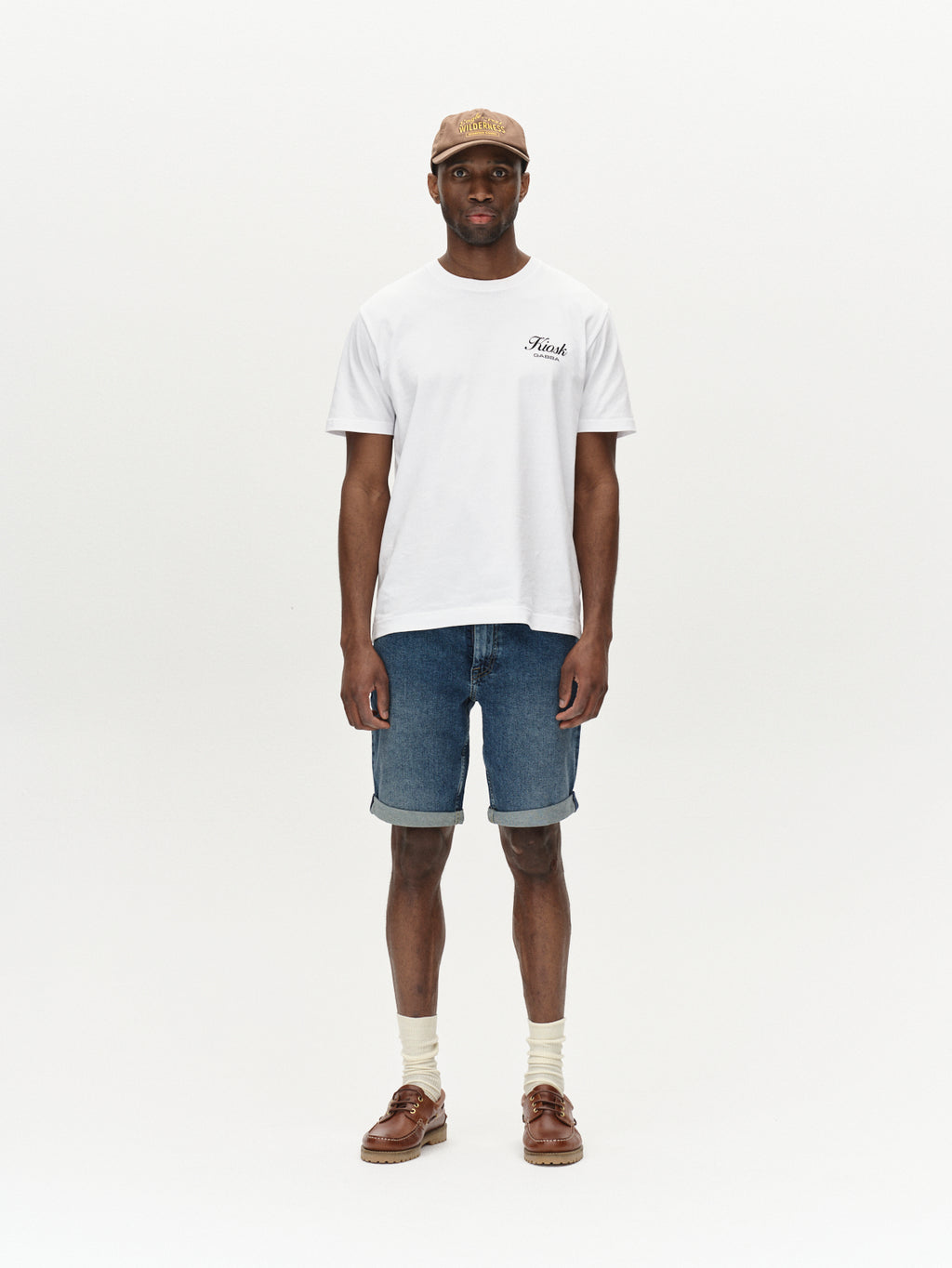 A person stands against a plain background wearing a beige cap, white Rich T-shirt, Gabbas Markus 5poc dark blue denim shorts for a relaxed vibe, beige socks, and brown shoes.