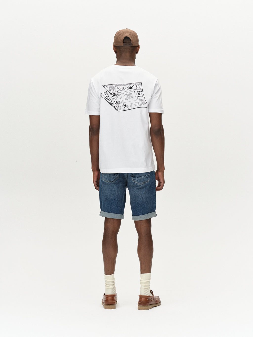 A man in a white T-shirt with a back graphic wears Gabbas Markus 5poc - Dark Blue Denim, offering comfort, along with brown shoes, a brown cap, and socks. He stands against a plain white background.