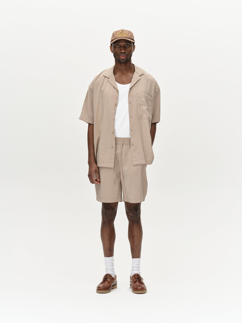 Against a plain background, a person sports the Gabba Jens Over Resort Yari SS in Sand Greige with matching shorts and a white undershirt. Paired with a beige baseball cap, white socks, and brown shoes, the outfit achieves a relaxed yet coordinated look.
